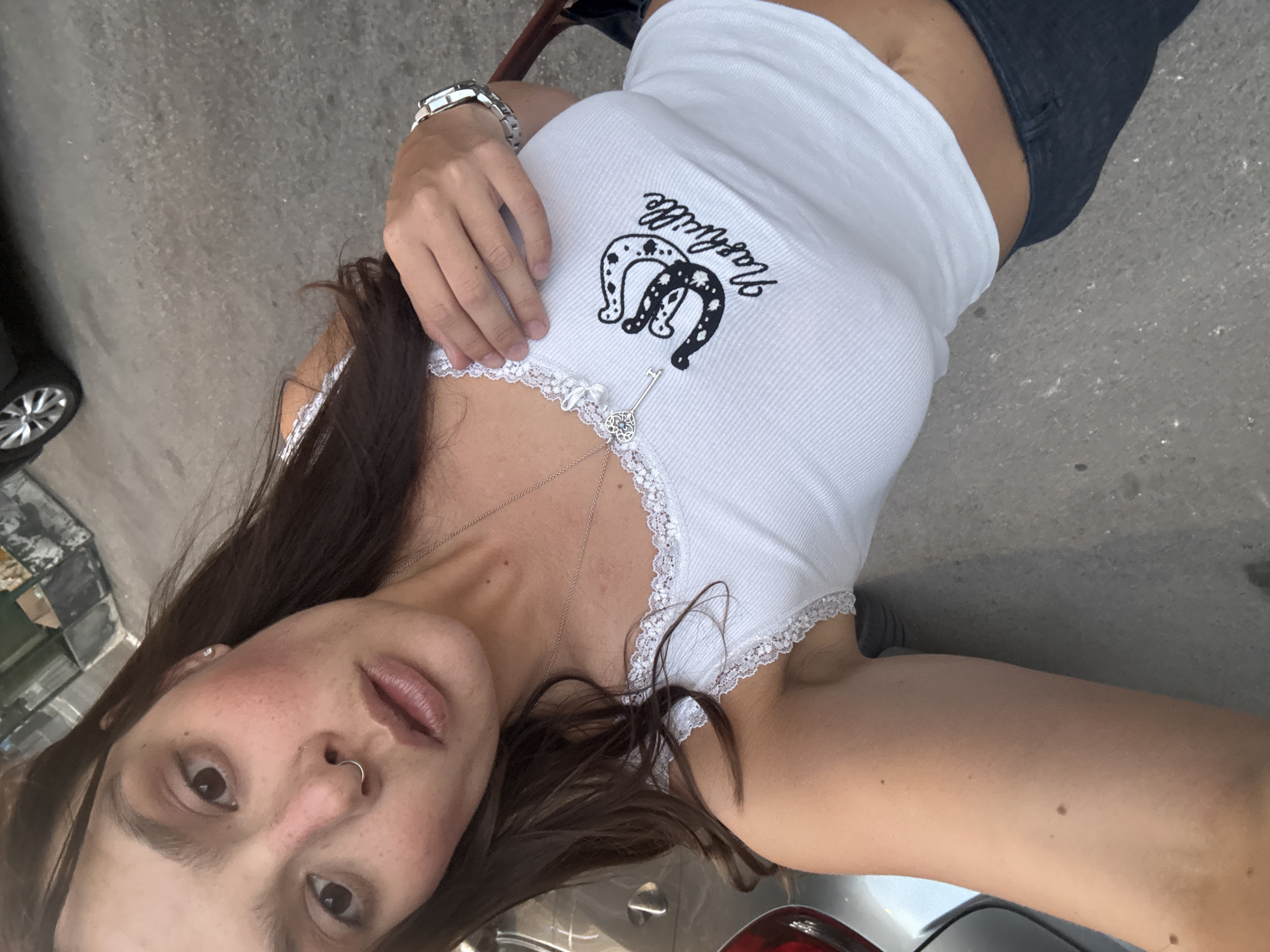 A young woman is taking a selfie outdoors, leaning against a car. She is wearing a white lace-trimmed tank top with a horseshoe and "Nashville" graphic on it, and dark denim jeans. She has a nose ring and is wearing a silver necklace with a key pendant and a watch on her left wrist. The background appears to be an urban street with a paved surface, a car tire, and some debris. There are no specific landmarks visible that would indicate the location is Tel-Aviv, Israel.