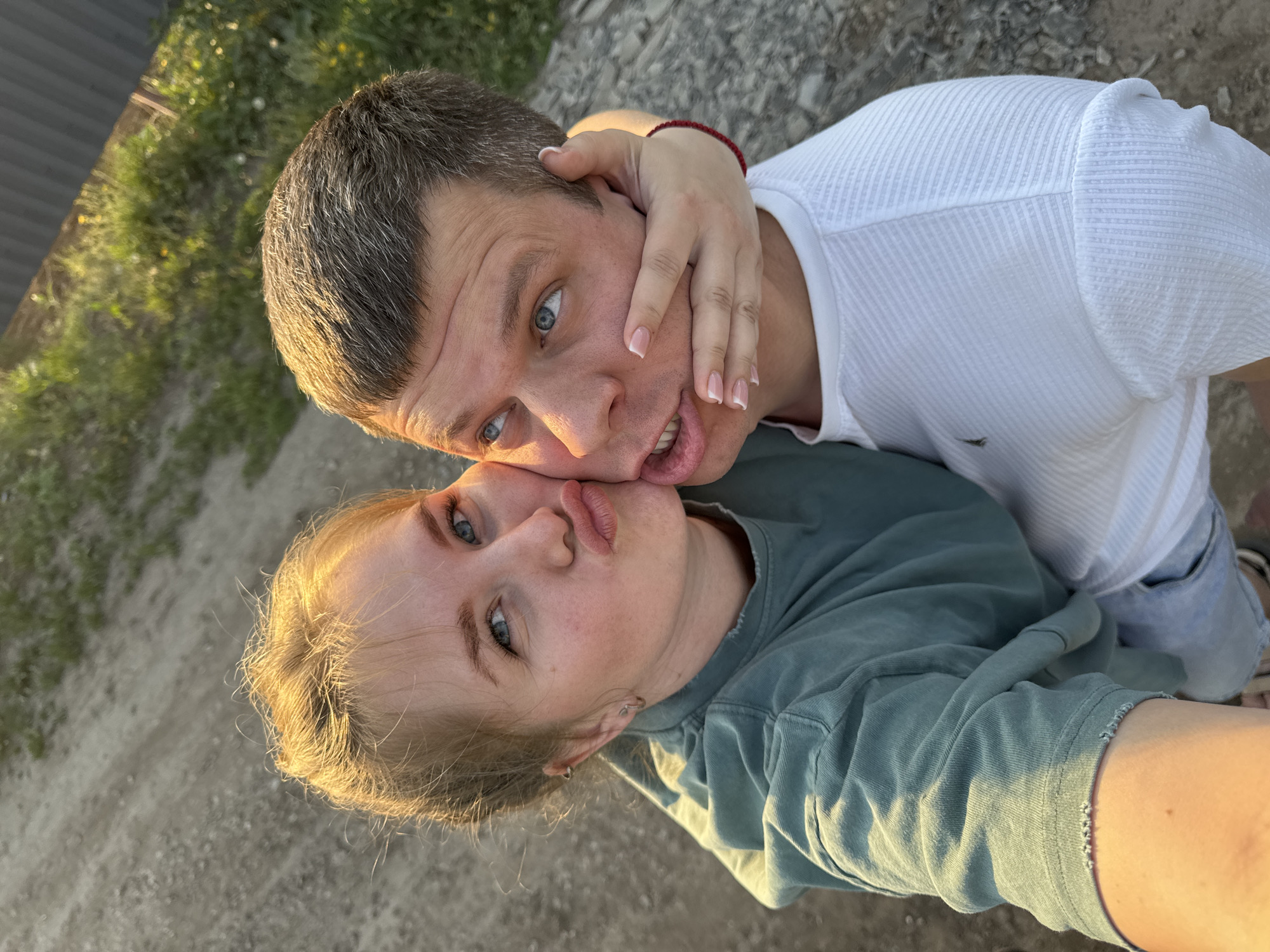 A man and a woman are posing for a selfie. The man has short, graying hair, blue eyes, and is wearing a white ribbed shirt. His mouth is open slightly. A hand with manicured nails is holding his face. The woman is wearing a green shirt. Both individuals have fair skin and blue eyes. The background consists of a gravelly surface, grass, and a fence. The photo appears to be taken outdoors in Горный, Russia. The image is slightly rotated counterclockwise.