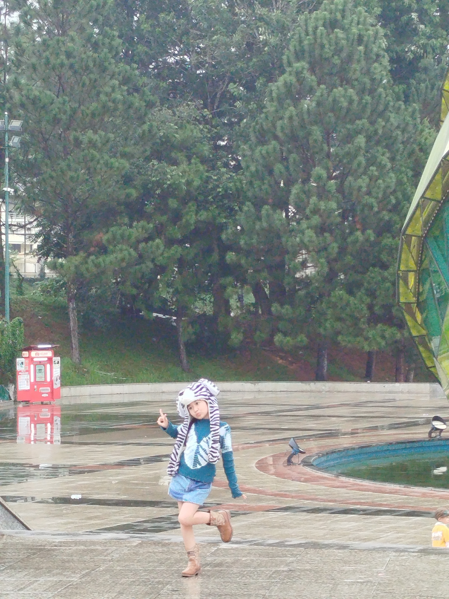 A child in a blue sweater, denim shorts, brown boots, and a zebra-striped hat with ears is posing on a wet, paved surface. The child is standing on one leg with the other raised, making a peace sign with one hand. The background features tall evergreen trees, part of a modern-looking building or structure with green and yellow accents, and a red box possibly containing public access devices. The scene appears to be a public square or park in Da Lat, Vietnam, based on the building design and the surrounding vegetation. A small portion of another child wearing a yellow shirt is visible in the lower right corner of the photo.