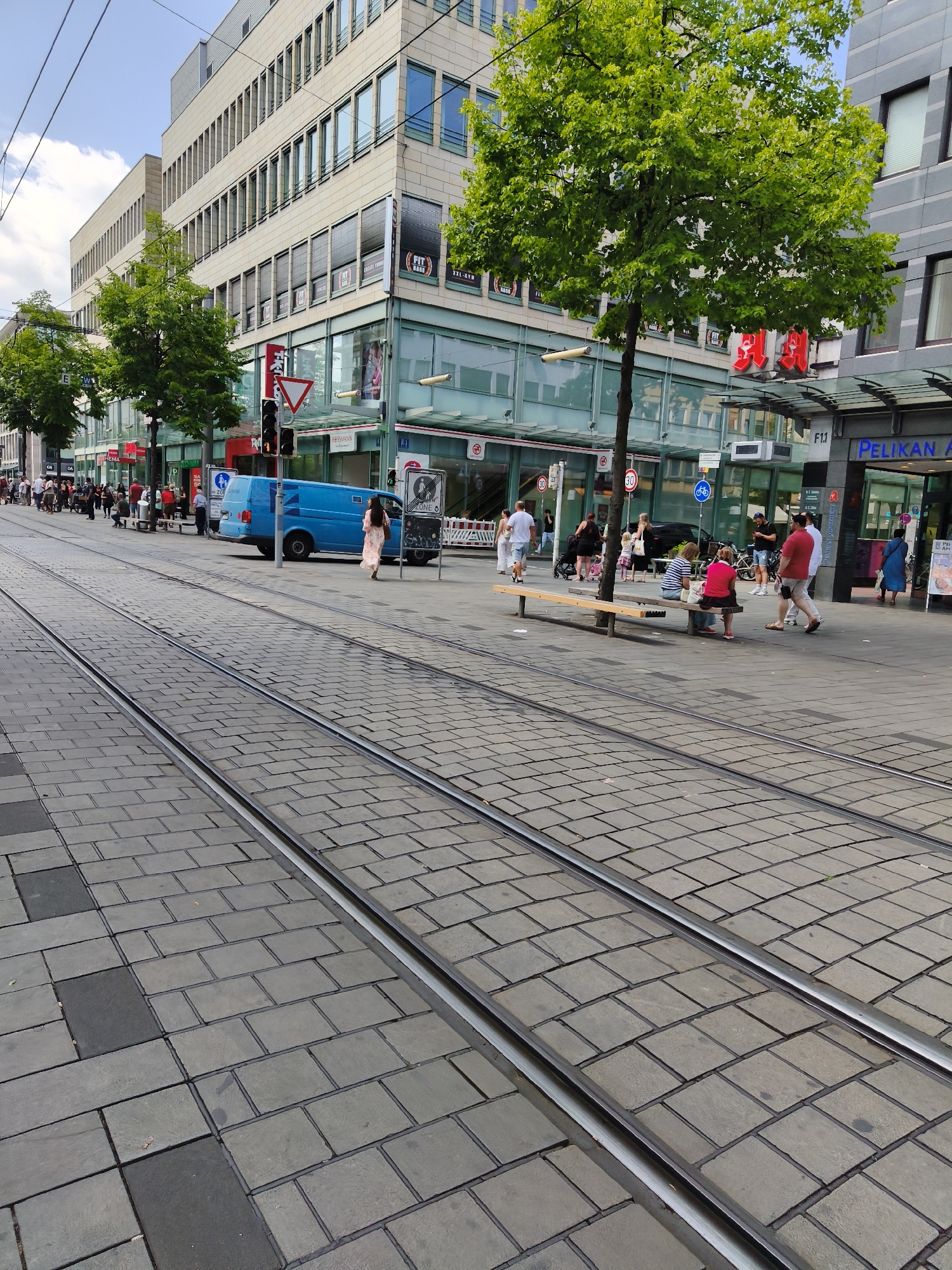 This is a street-level, eye-level, full shot of a city street in Mannheim, Germany, featuring tram tracks in the foreground and a modern building with storefronts in the background. The tram tracks are made of metal and run parallel to each other, embedded in a grey, paved surface composed of rectangular blocks.
Several people are visible on the sidewalk. On the left side, a line of people are waiting, some standing and some sitting on a long, slatted bench. A blue van is parked on the street to the right of the tram tracks. Further down the street, on the right side, several more individuals are walking or standing.
The building behind the sidewalk is a multi-story structure with a beige facade. Its windows are rectangular and covered with blinds. The ground floor of the building features large glass storefronts with visible signage. One sign clearly reads "PELIKAN APOTHEKE". Several trees with lush green foliage are planted along the sidewalk, providing shade. The sky is partly cloudy. The scene depicts a typical urban environment with a focus on pedestrian and public transport infrastructure.