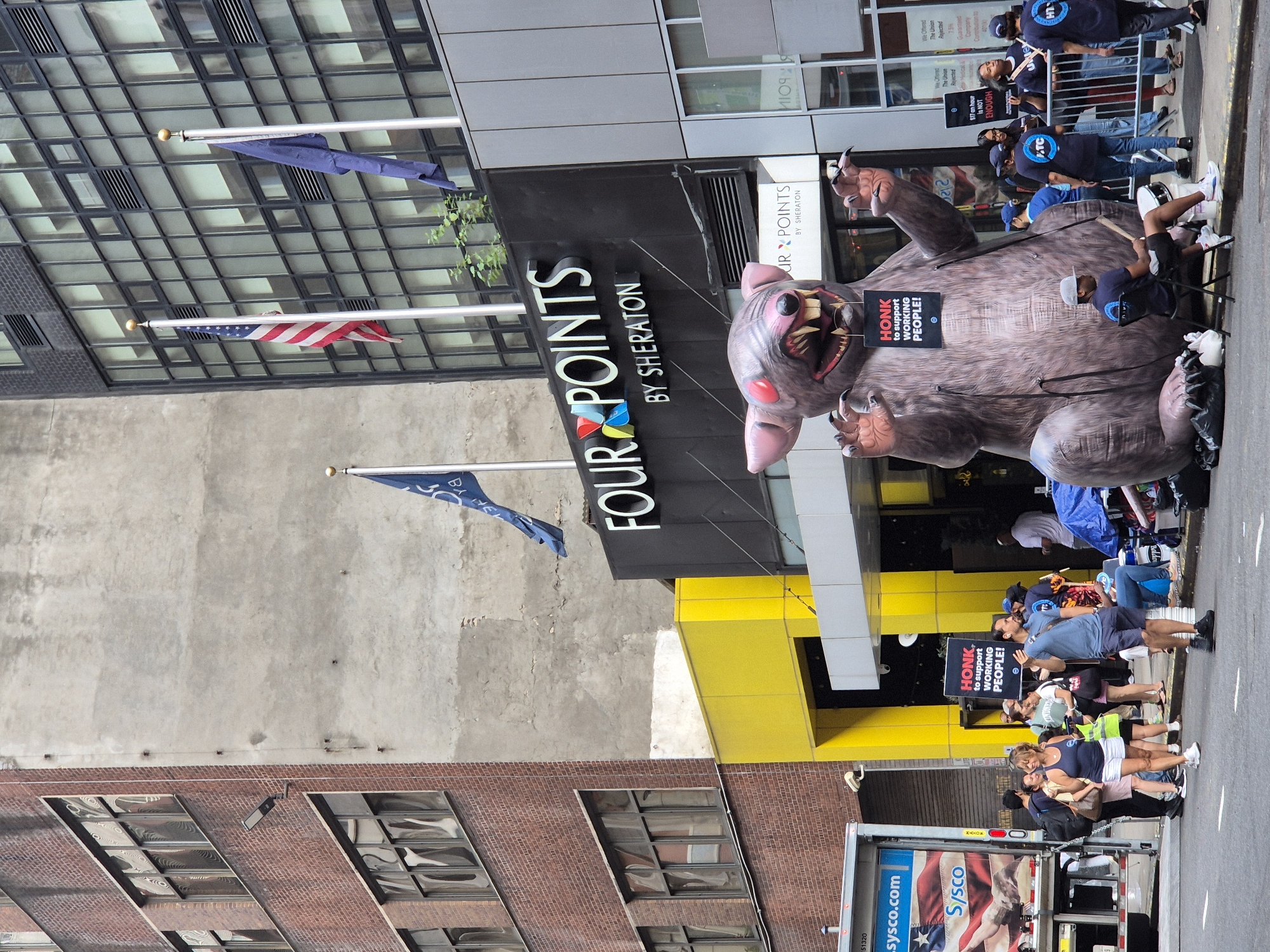 The image depicts a street scene in New York City, USA, outside the Four Points by Sheraton hotel. A large inflatable rat, a common symbol used in labor disputes, is prominently displayed on the sidewalk. The rat is holding a sign that reads "HONK to support WORKING PEOPLE!". A group of people are gathered around the inflatable rat, some holding signs and appearing to be protesting or showing support. One person is playing a drum, adding to the atmosphere of a demonstration. The background shows the hotel's facade with its signage, and adjacent buildings, including a brick building and a modern glass-fronted building. A Sysco truck is parked on the street to the left of the scene. Several individuals are visible in the image, engaged in various actions such as standing, walking, and holding signs. The overall impression is that of a public demonstration related to labor issues.