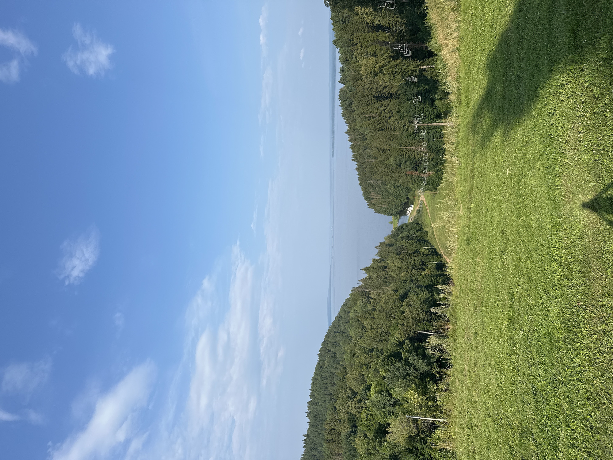 The image depicts a scenic vista overlooking a body of water, likely a lake or reservoir, bordered by dense green forests. In the foreground, a grassy slope is visible, with a shadow cast across it, suggesting a person or object is present just outside the frame. A ski lift with empty chairs stretches across the middle ground, ascending the wooded hillside. The sky is a clear blue with scattered white clouds. The location is identified as Galevo, Russia.