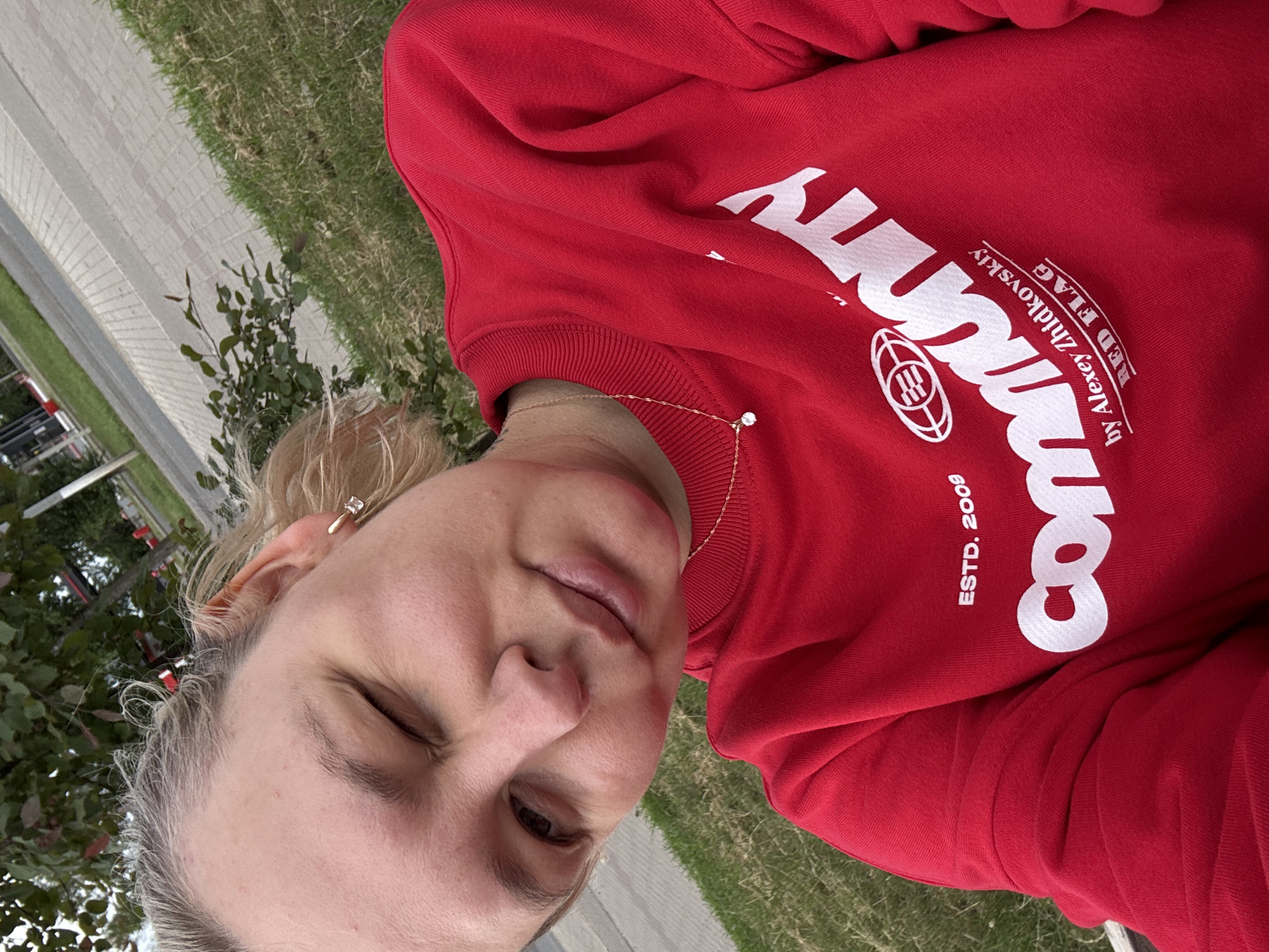 A woman with blonde hair, wearing a red sweatshirt and a gold necklace, winks at the camera. The sweatshirt has white text that reads "Community" along with "ESTD. 2009", "by Alexey Zhidkovskiy", and "RED FLAG". The background shows a paved walkway, green grass, and trees, suggesting an outdoor urban setting. The image was taken in Chaikovsky, Russia.