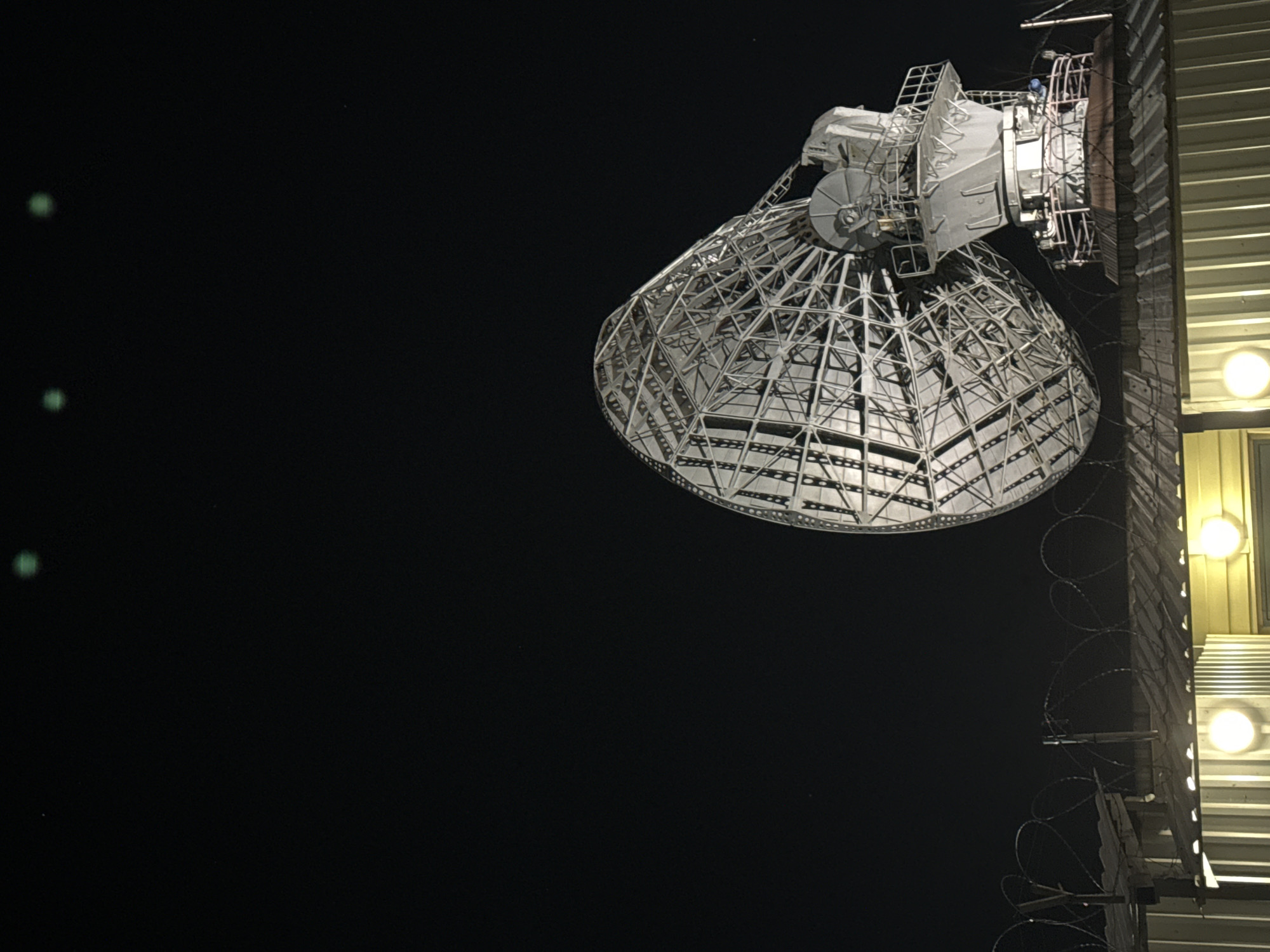 The image shows a large radio telescope antenna in Krasnoyarsk, Russia, illuminated against a night sky. The antenna is a complex structure of metal framework, dish-shaped, and mounted on a tower or building. Barbed wire runs along the edge of the building, suggesting security measures. Several lights are visible on the building. Three faint green orbs are visible in the upper left, possibly related to light sources or atmospheric phenomena.