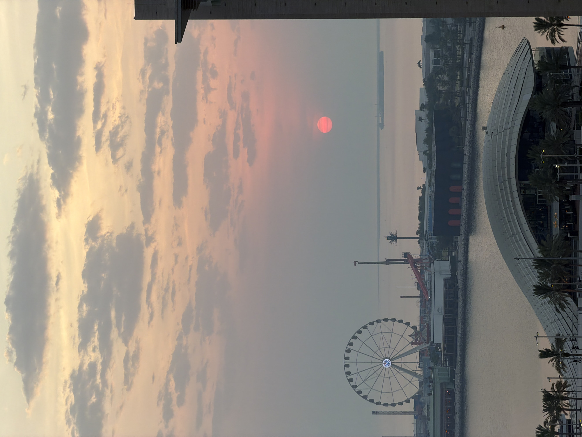 The image depicts a coastal city skyline at sunset, likely Lusail, Qatar. The sun, a vibrant pinkish-red orb, is low on the horizon, partially obscured by a hazy sky filled with scattered clouds painted in soft hues of peach and lavender. In the foreground, a body of water reflects the muted colors of the sky. On the waterfront, a large Ferris wheel stands prominently, its structure silhouetted against the twilight. Several buildings, some with dark facades and others with lighter structures, line the shore, interspersed with palm trees. A large, modern building with a distinctive curved roof and glass facade is visible in the lower part of the frame, featuring illuminated windows and surrounding palm trees. In the distance, a large cargo ship sails on the calm sea. The overall atmosphere is serene and tranquil, characteristic of a coastal sunset.