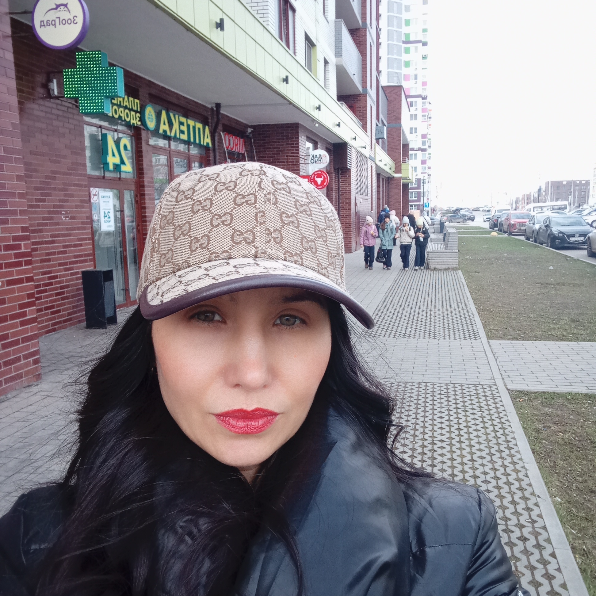 The image is a selfie featuring a woman in the foreground on a city sidewalk, with an urban streetscape behind her. The woman, who has long dark hair and red lipstick, is wearing a light brown baseball cap with a patterned logo (resembling Gucci's "GG") and a black puffy jacket. She looks directly at the camera with a neutral expression.
Behind her, on the left, is a brick building with ground-floor commercial units. Several signs are visible, displaying Cyrillic text that appears reversed, suggesting the image is a mirror selfie. A large green cross sign indicates a pharmacy. Reversed text clearly visible on the pharmacy storefront includes "АПТЕКА" (APTEKA - Pharmacy) and a "No Smoking" sign. Above the pharmacy, a round purple sign with a cat logo also shows reversed Cyrillic text, which would read "ЗООТЕР" (ZOOSTORE) when corrected. Another small white circular sign has a bull logo and some inverted text.
Further down the sidewalk, several individuals, including children dressed in warm jackets and hats, are walking away from the camera. The sidewalk features a patterned paving design, and a strip of sparse grass runs alongside the street. The street itself is lined with numerous parked cars. In the background, tall, multi-story residential buildings with distinct colorful panels are visible against a bright, overcast sky. The overall scene depicts a cool, daytime environment in an urban area, consistent with the provided location context of Izhevsk, Russia.
