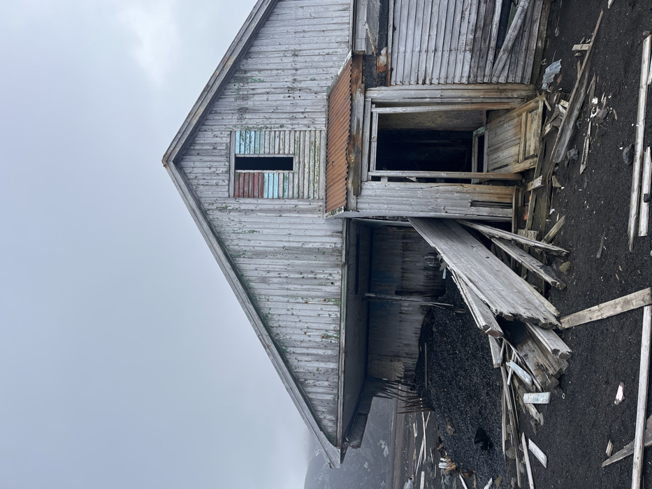 A dilapidated wooden structure stands against a cloudy sky. The building's weathered facade exhibits peeling paint and decaying timber. A partially obscured window with remnants of blue and red paint sits near the roofline. A gaping doorway leads to darkness, framed by rotting wood. Debris litters the ground, consisting of shattered planks and scattered material, suggesting abandonment and neglect. The earth's dark tone contrasts with the structure's faded color.