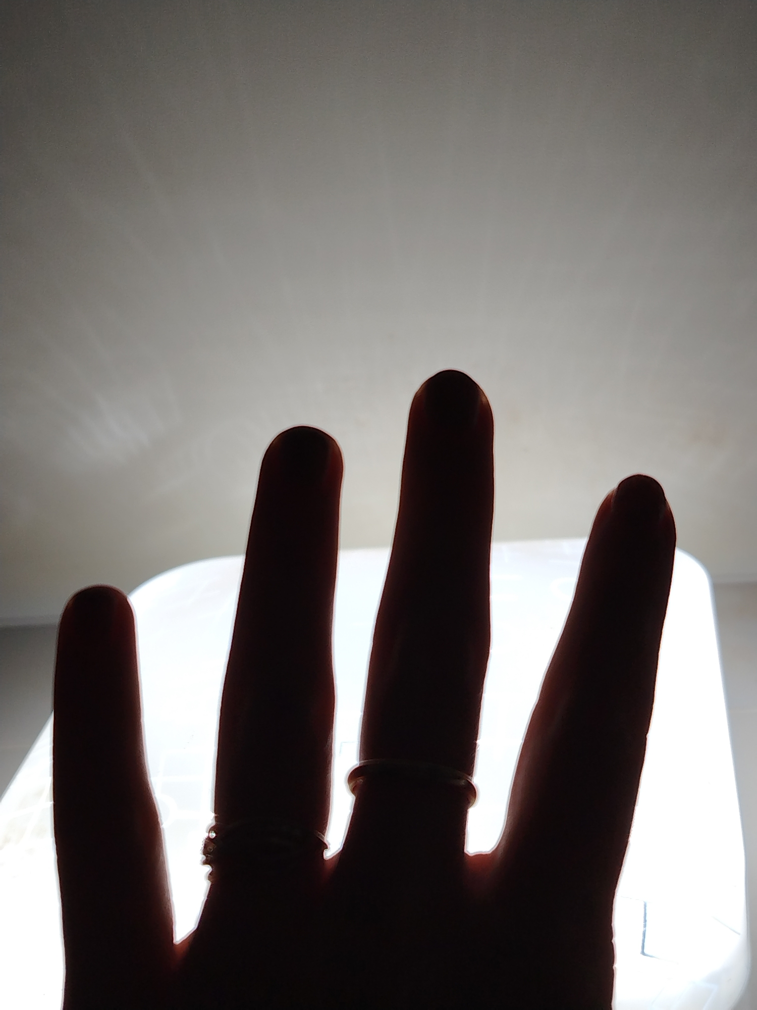 A close-up, silhouetted view of a hand against a bright light source. Four fingers are visible, partially obscuring the light. There are rings on two of the fingers. The background is a blurred light source with some details suggestive of a room. No specific details provide information related to the city of Izhevsk, Russia.