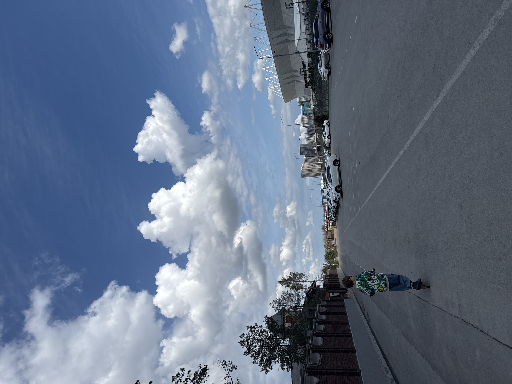 A child is riding a scooter on a paved area in Rostov-on-Don, Russia. Cars are parked along the side. A building is visible in the background, with a blue sky and clouds above.