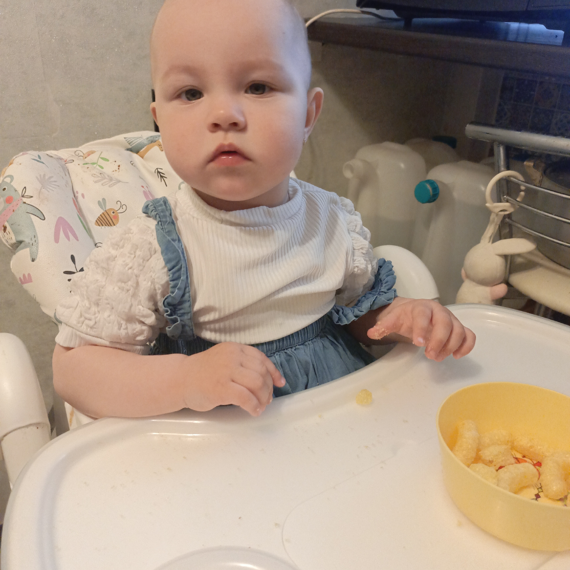 A baby with fair skin and dark eyes sits in a white high chair, looking directly at the camera with a neutral expression. The baby wears a white, ribbed long-sleeved shirt with ruffled shoulders and a light blue denim-like overall dress with ruffled shoulder straps. Her high chair features a patterned fabric insert with illustrations of koalas, bees, and leaves.
On the white tray of the high chair, a yellow plastic bowl contains several small, light-colored, irregular-shaped food pieces, possibly puffed snacks or soft fruit. A few similar food pieces are scattered on the tray near the bowl and on the tray to the left. The baby's right hand has some food residue on it.
The background is an indoor setting, likely a kitchen or dining area. To the left, a light-colored wall is visible. To the right, various items are stacked or placed on a surface. These include several translucent white plastic jugs with blue or green caps, a small white bunny figurine or toy hanging from a metal rack, and a dark shelf or countertop above. A dark electronic device is partially visible at the top right. The overall lighting suggests an indoor, daytime scene. There is no visible text in the image.