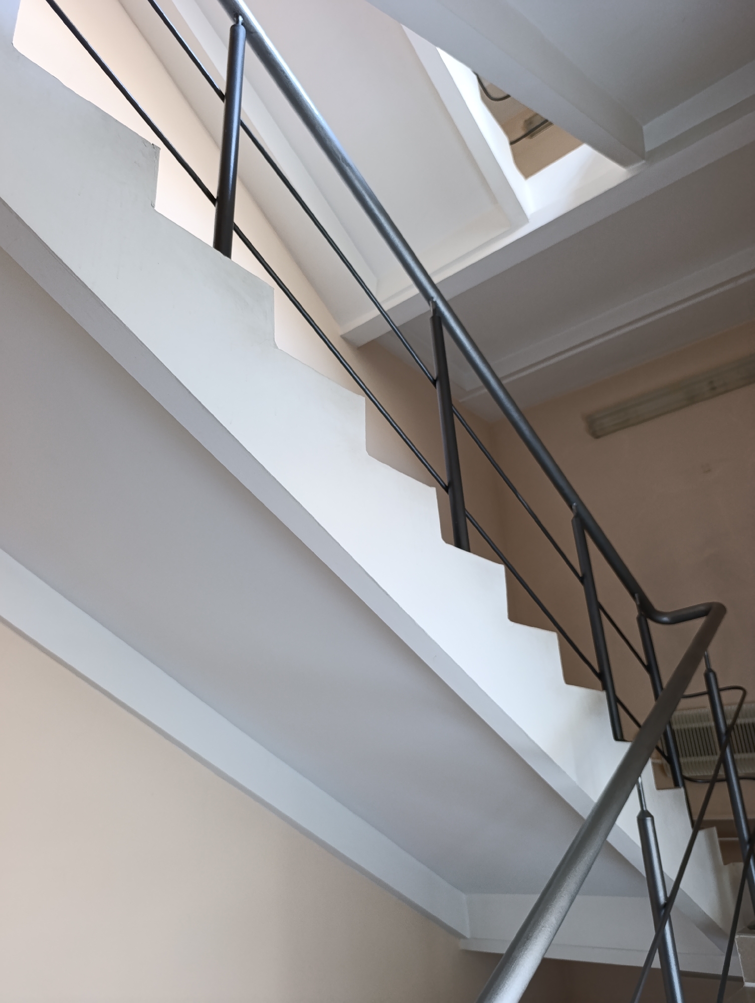 The image shows an interior view of a staircase with a metal railing. The staircase has white risers and treads, with a light-colored wall behind it. The railing is dark gray and consists of a top handrail and several vertical balusters. The scene appears to be located in an indoor public space, possibly a building in the city of Dubăsari town council, Moldova, given the context provided. The lighting is somewhat dim, with a fluorescent light fixture visible on the wall in the background. There are no people or animals visible in the image. The composition is a low-angle shot looking up the staircase.