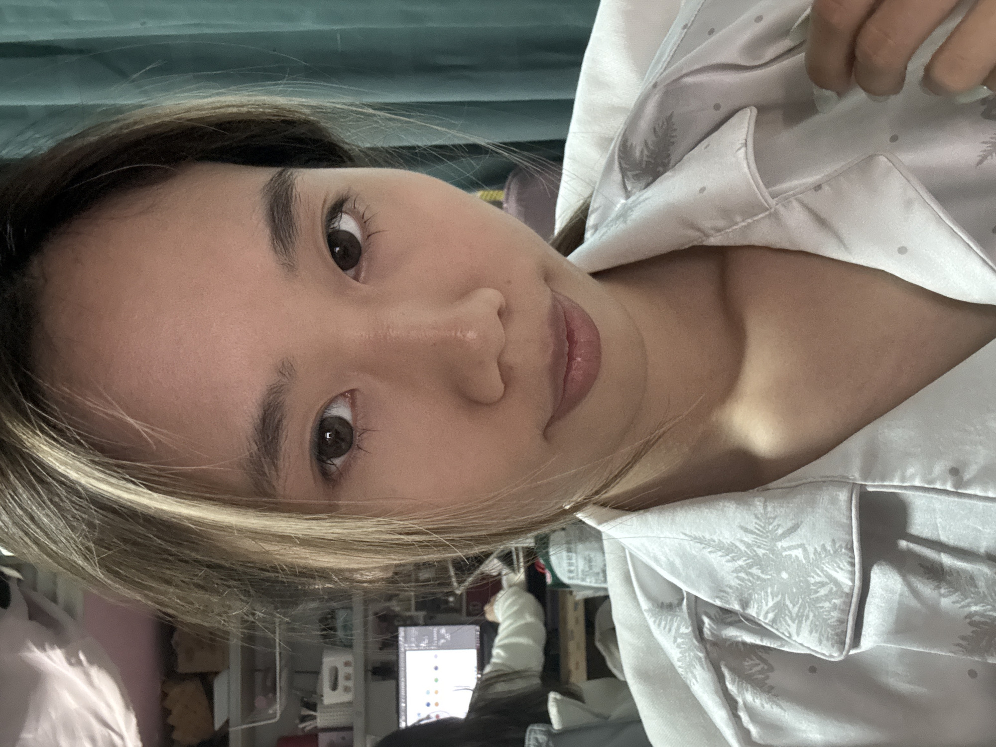 The image shows a person with brown eyes and light brown and blonde hair, wearing a light-colored button-down shirt with a faint pattern of pine trees and dots. The person is lying down, with the camera positioned at an angle to their face.
Behind the person, there is a cluttered area with a computer monitor displaying colorful dots, shelves with various objects, and a white chair. A green curtain is visible in the background.
There is no information to support the Nan'an District location.