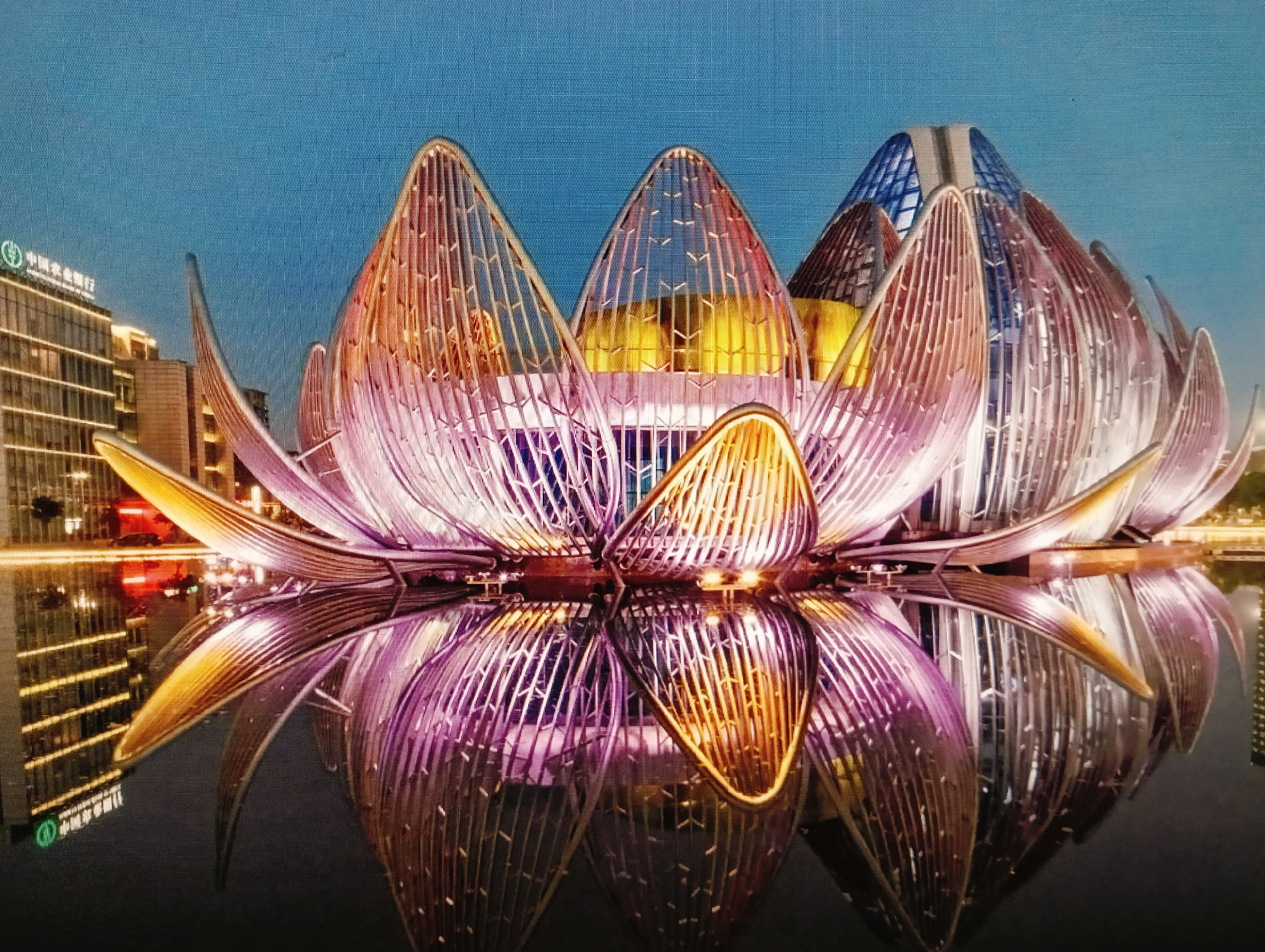 The image displays a striking lotus-shaped building, illuminated at dusk. The structure, composed of translucent petals that glow with pink and gold hues, is reflected in the dark water below. Behind the lotus, a modern building with glowing windows is visible, suggesting an urban setting. The sky above is a deep blue, indicative of twilight. There are no people visible in the scene. The image is a photograph of the "Lotus Plaza" in the city of Slutsk, Belarus.