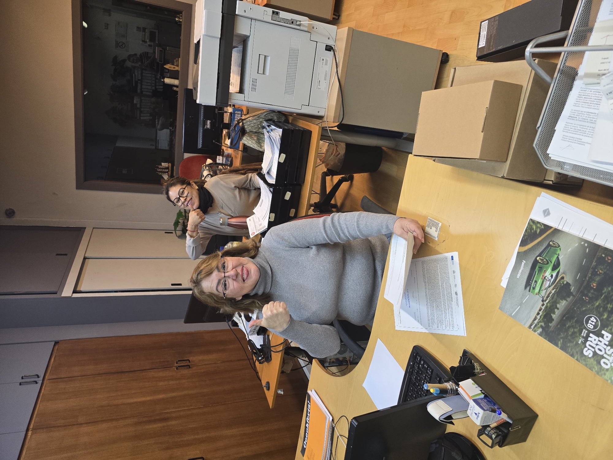 The image displays an indoor office environment, captured from an overhead perspective and rotated ninety degrees clockwise. Two women are visible, seated at light wooden desks, both smiling and engaging with the camera.
In the foreground, a woman with shoulder-length, reddish-brown hair and glasses is seated at a desk, wearing a grey turtleneck sweater. She is holding papers in one hand and enthusiastically giving a thumbs-up with the other, her gaze fixed on the camera with a friendly smile. Her desk is cluttered with typical office items, including a black laptop, a keyboard, a pen holder with various writing tools, and a small white box that appears to be medical, with "Valdis" partially visible. A mesh paper tray overflows with documents, and next to it lies a magazine or brochure featuring a green sports car on a winding road, with visible text including "Fascinaçión", "418", and "PHO RUS". Documents on the desk also show what resembles an EU logo and text in a Romance language. An orange folder on the desk has the word "Oooohferta!" printed on it.
Behind her, a second woman with dark hair and glasses, dressed in a light-colored top and a black neck scarf, is also seated at a desk, looking towards the camera with a gentle smile. In the background, a large grey office photocopier/printer stands prominently on a desk. Further back, a large glass partition or window reflects the office interior, suggesting an adjacent space. The walls feature various wooden cabinets, some with dark brown doors and others with lighter grey or beige panels. Several cardboard boxes are stacked on a nearby desk.
The overall atmosphere is positive and welcoming, conveyed by the women's smiles and the thumbs-up gesture. The setting is a functional, busy office space, consistent with workplaces found in urban environments like Barcelona, Spain.