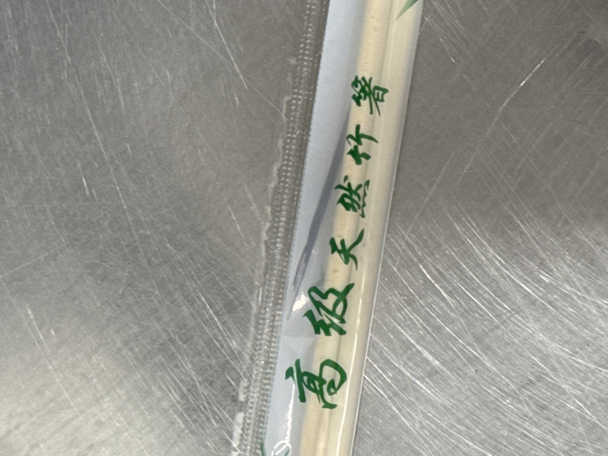 The image shows a close-up of a pair of chopsticks in plastic packaging. The chopsticks are made of wood and have green Chinese characters printed on them. The background is a metallic surface with visible scratches. The image appears to have been taken in a kitchen or food service environment. There is no indication of the location in the city of Mariupol, Ukraine, in the provided image.