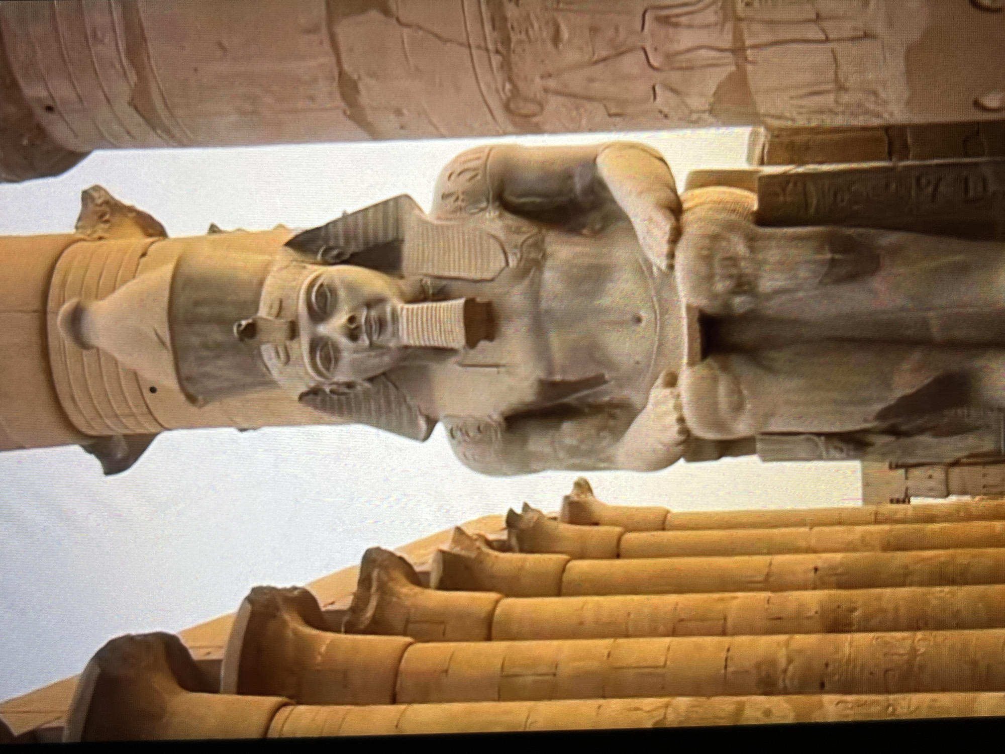 The image shows an Egyptian statue, possibly of Ramses II, in the Luxor Temple complex, Egypt, not Tinaquillo, Venezuela. The statue stands among columns and carved stonework typical of ancient Egyptian architecture. The statue wears traditional pharaonic headdress and attire. The bottom of the statue has hieroglyphics and a row of columns, typical of Egyptian buildings. The perspective indicates the image was taken from below, looking up at the statue and surrounding structure.