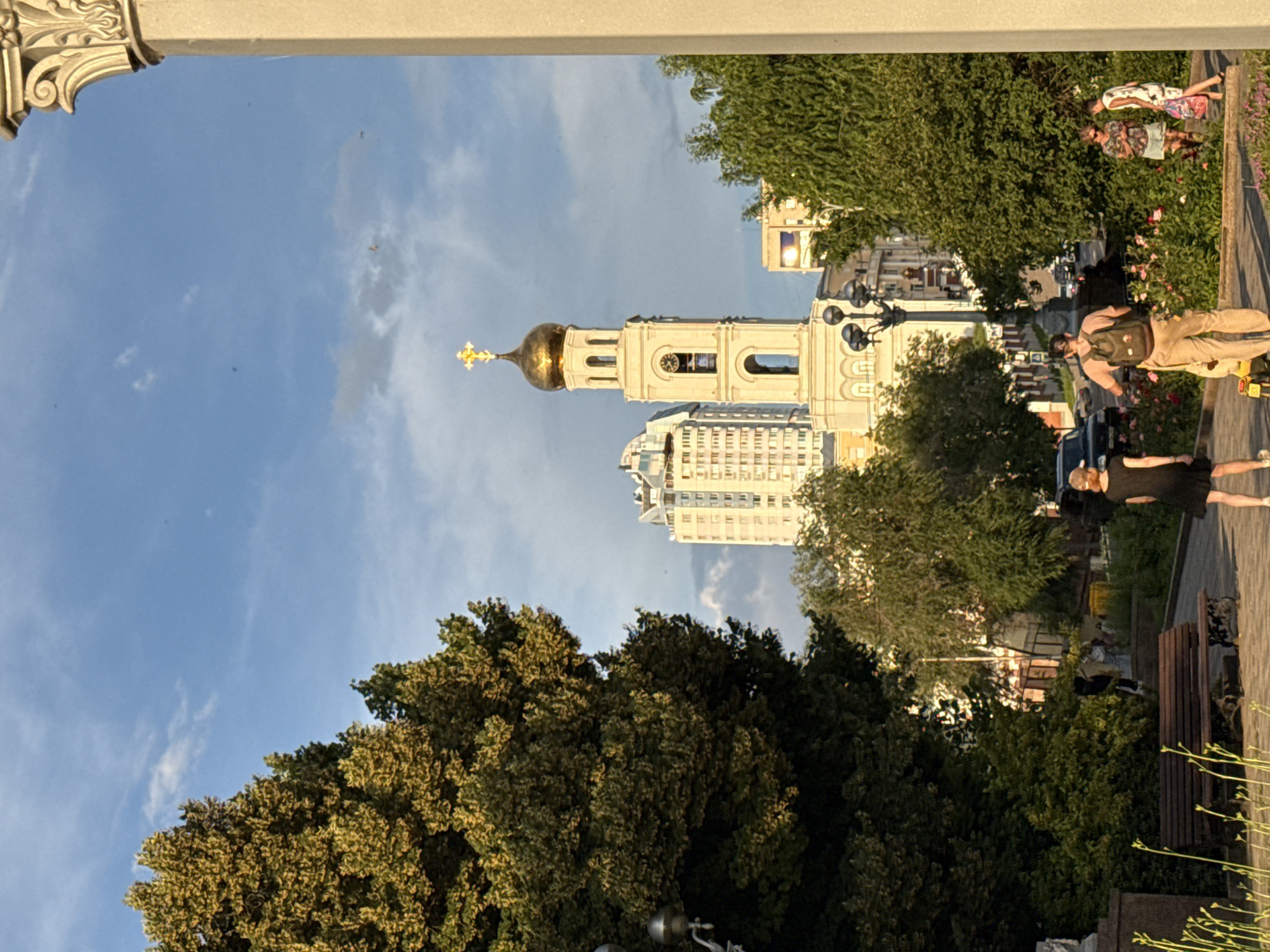 The image depicts a street scene in Samara, Russia, featuring a prominent bell tower with a golden dome and a cross, likely part of a church. The tower is framed by lush green trees and a modern high-rise building in the background. In the foreground, several people are visible. On the left, a person in a black dress walks away from the camera. In the middle, a person with a backpack on a yellow cart is facing away. To the right, two women are standing and interacting. The scene appears to be set in the late afternoon or early evening, with warm sunlight illuminating parts of the buildings and trees. The sky is blue with scattered clouds.