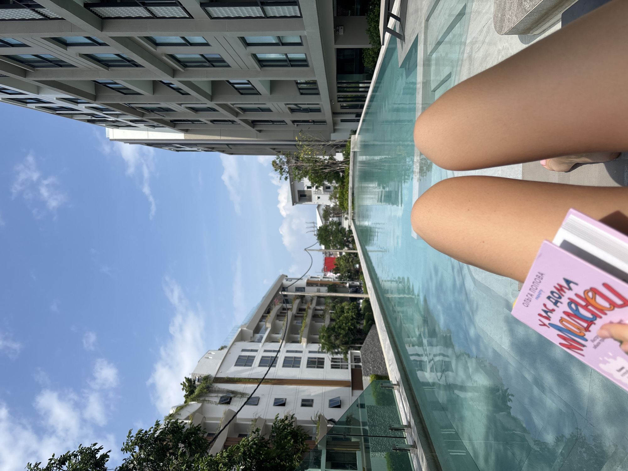 The image shows a view of a person sitting poolside in Choeng Thale, Thailand. The person's legs are visible in the foreground, holding a pink book. The pool water reflects the surrounding buildings and the blue sky. Two buildings of different styles are visible across from each other. Trees are also growing next to one building.