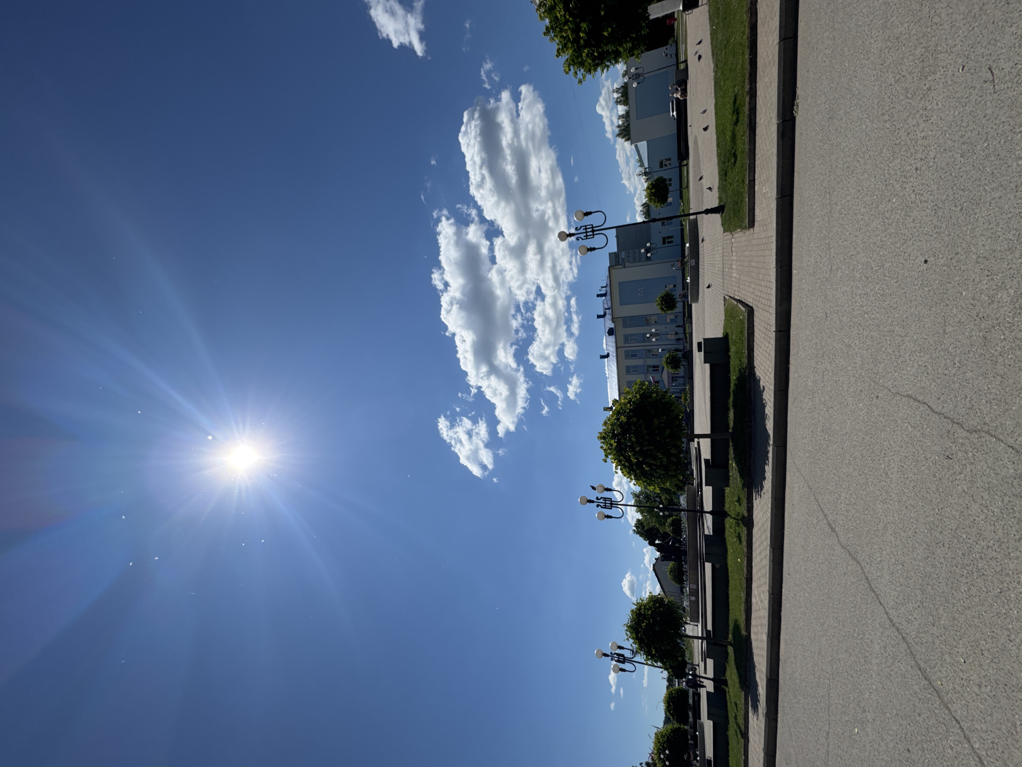 The image depicts a bright, sunny day in a city square, likely in Chaikovsky, Russia, given the visible Cyrillic signage on the blue and white building. The sun is prominently featured in the upper left quadrant, casting lens flares and illuminating a clear blue sky dotted with fluffy white clouds.
In the foreground, a paved road with visible cracks transitions into a walkway made of interlocking bricks. This walkway borders a manicured lawn area with several small, round trees. Two ornate streetlights with multiple globes stand in this green space.
Further back, a large, modern building with blue and white facades dominates the background. Cyrillic text, including what appears to be "КУЛЬТУРНЫЙ ЦЕНТР" (Cultural Center) and the letter "М," is visible on its side. A fountain is situated in front of the building. Scattered throughout the square are several pigeons. A few people are visible walking in the distance, and a dark-colored statue or monument is present in the middle ground. A parked white car is visible near the building on the right side of the frame. The overall impression is of a well-maintained public space on a pleasant day.