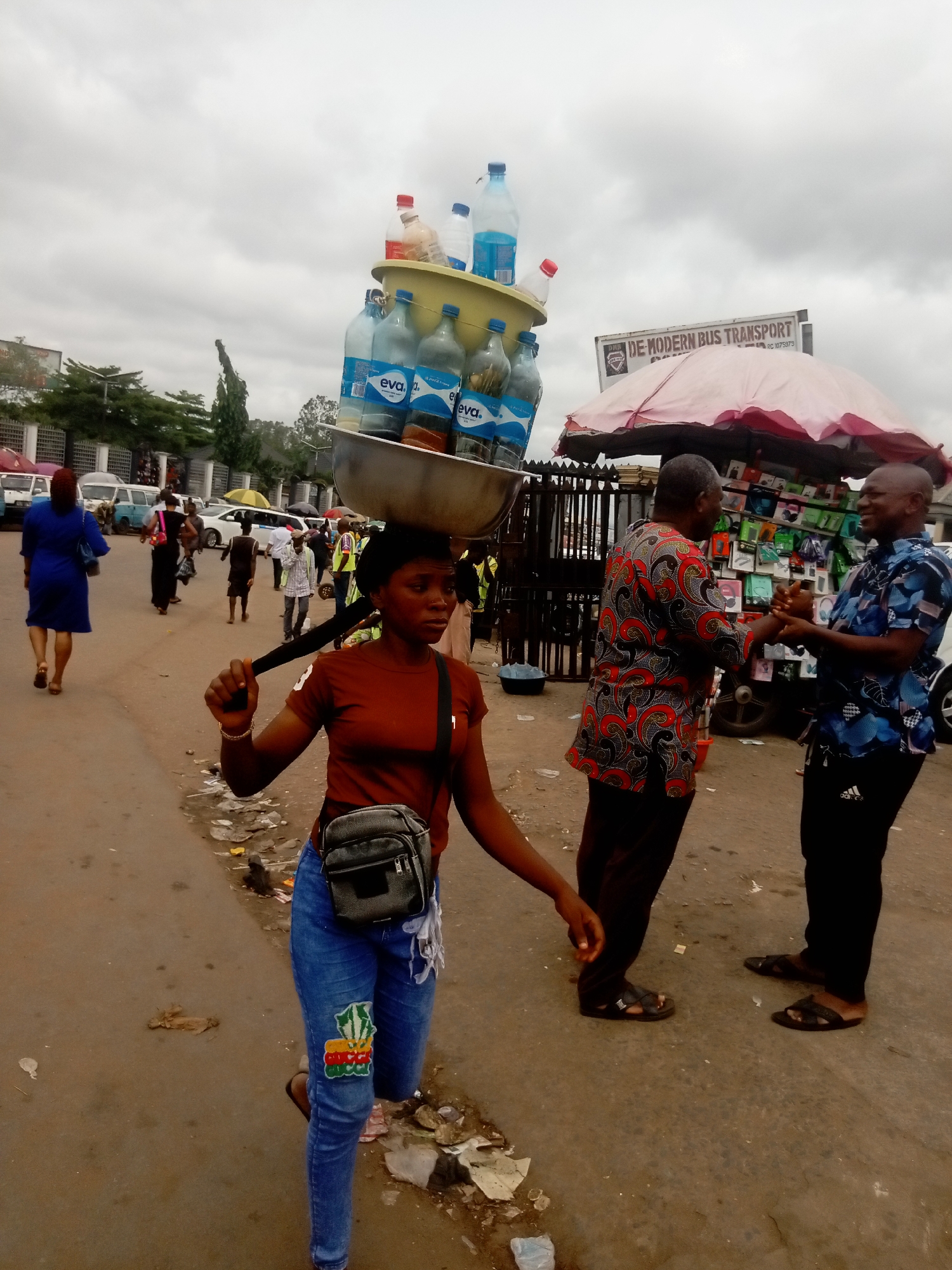 The image captures a street scene in Onitsha, Nigeria. A young woman, wearing a maroon t-shirt, blue jeans with a colorful patch, and a crossbody bag, balances a large metal bowl filled with bottled water on her head. She holds a black object in her hand, possibly for balance. To the right of the woman, two men engage in a handshake under an umbrella sheltering a display of phone accessories. A sign reads "DE MODERN BUS TRANSPORT". In the background, other pedestrians and vehicles are visible, suggesting a bustling urban environment under an overcast sky. The street appears worn, with litter scattered on the ground.