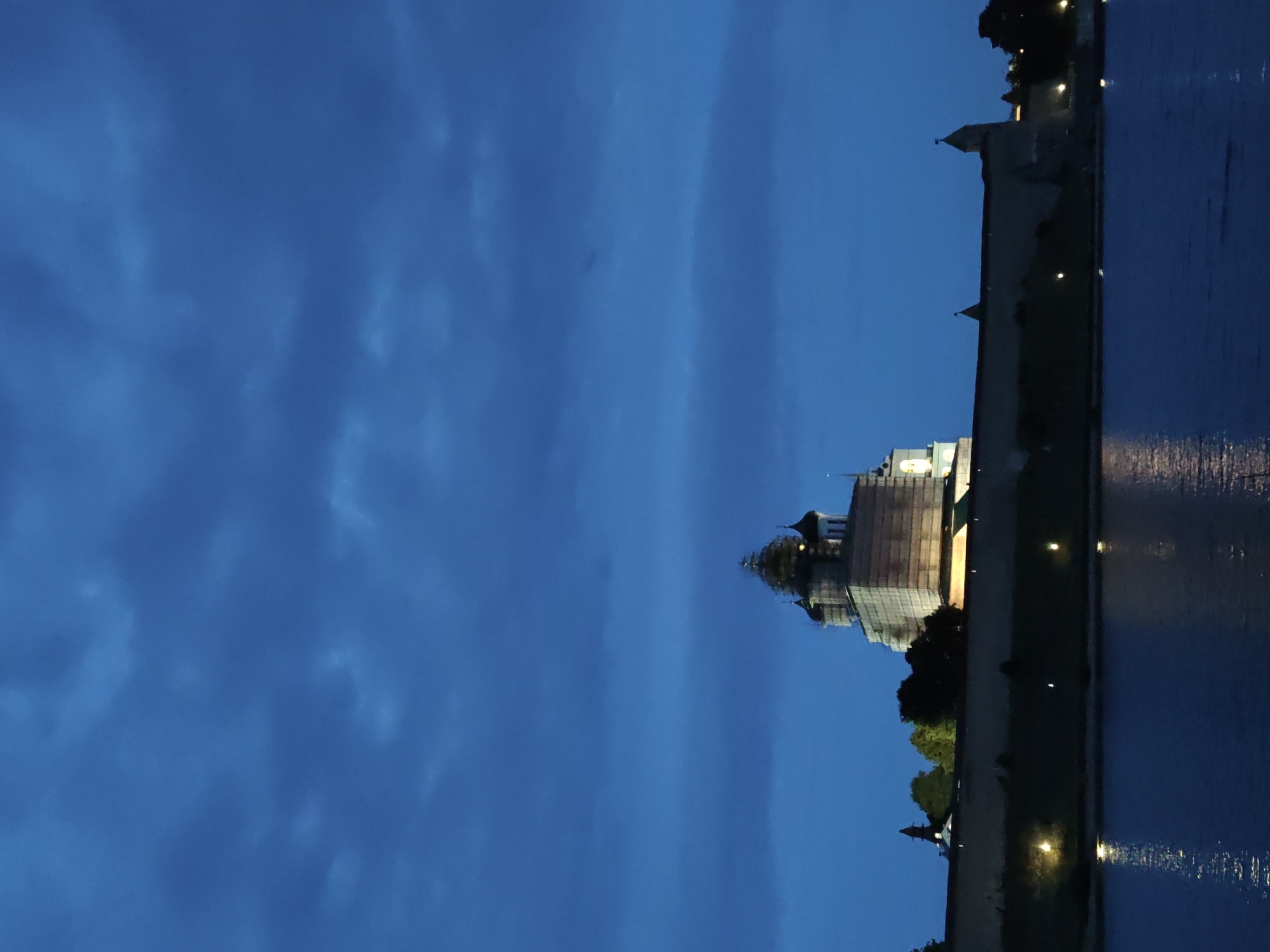 A low-angle, vertical shot depicts the Pskov Kremlin and its fortifications at dusk, illuminated by artificial lights. The sky is a deep, cloudy blue. The Kremlin, a large stone fortress with multiple towers and a prominent cathedral with onion domes, is the central focus. The cathedral appears to be undergoing renovations, as scaffolding is visible around its upper sections. A calm body of water, likely the Pskov River, occupies the bottom third of the image, reflecting the lights from the fortress and the twilight sky. The water's surface is mostly smooth, with gentle ripples. The lighting suggests it is after sunset, with the building and its surroundings lit for nighttime visibility.