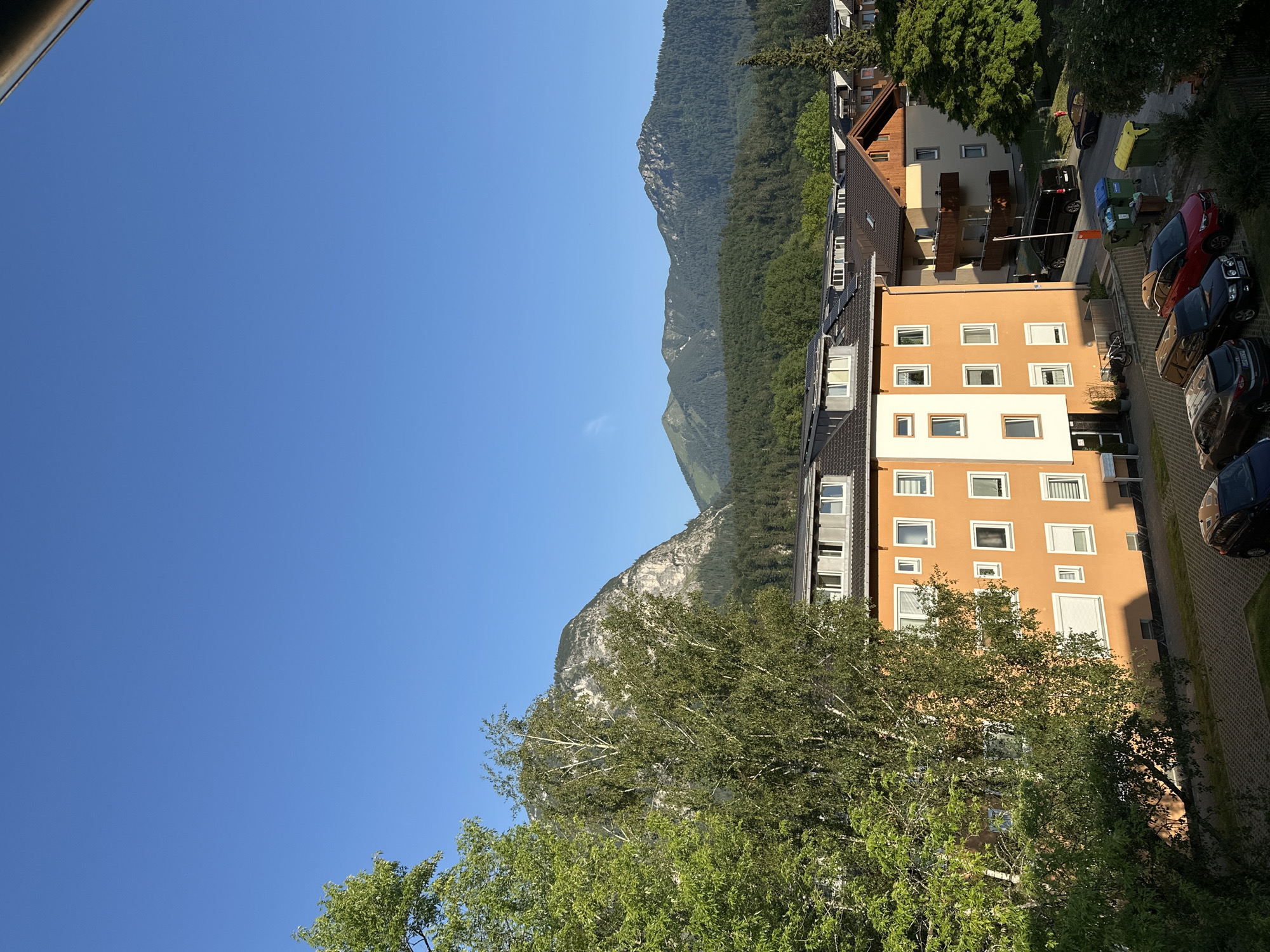 This image depicts a view of Garmisch-Partenkirchen, Germany, from an elevated vantage point. The scene is dominated by a bright, clear blue sky. In the foreground, a large deciduous tree with vibrant green leaves partially obscures the view of a multi-story apartment building. The building is painted a distinct orange color and features numerous windows with white frames. Adjacent to this building, other residential structures are visible, including one with a dark tiled roof and a wooden balcony. Below the buildings, a paved area serves as a parking lot where several cars are parked. Further in the background, a dense, dark green forest covers the slopes of mountains. The mountains themselves are rugged, with lighter colored rock faces and patches of snow, indicating a high altitude. The overall impression is one of a serene, daytime scene in a mountainous region.