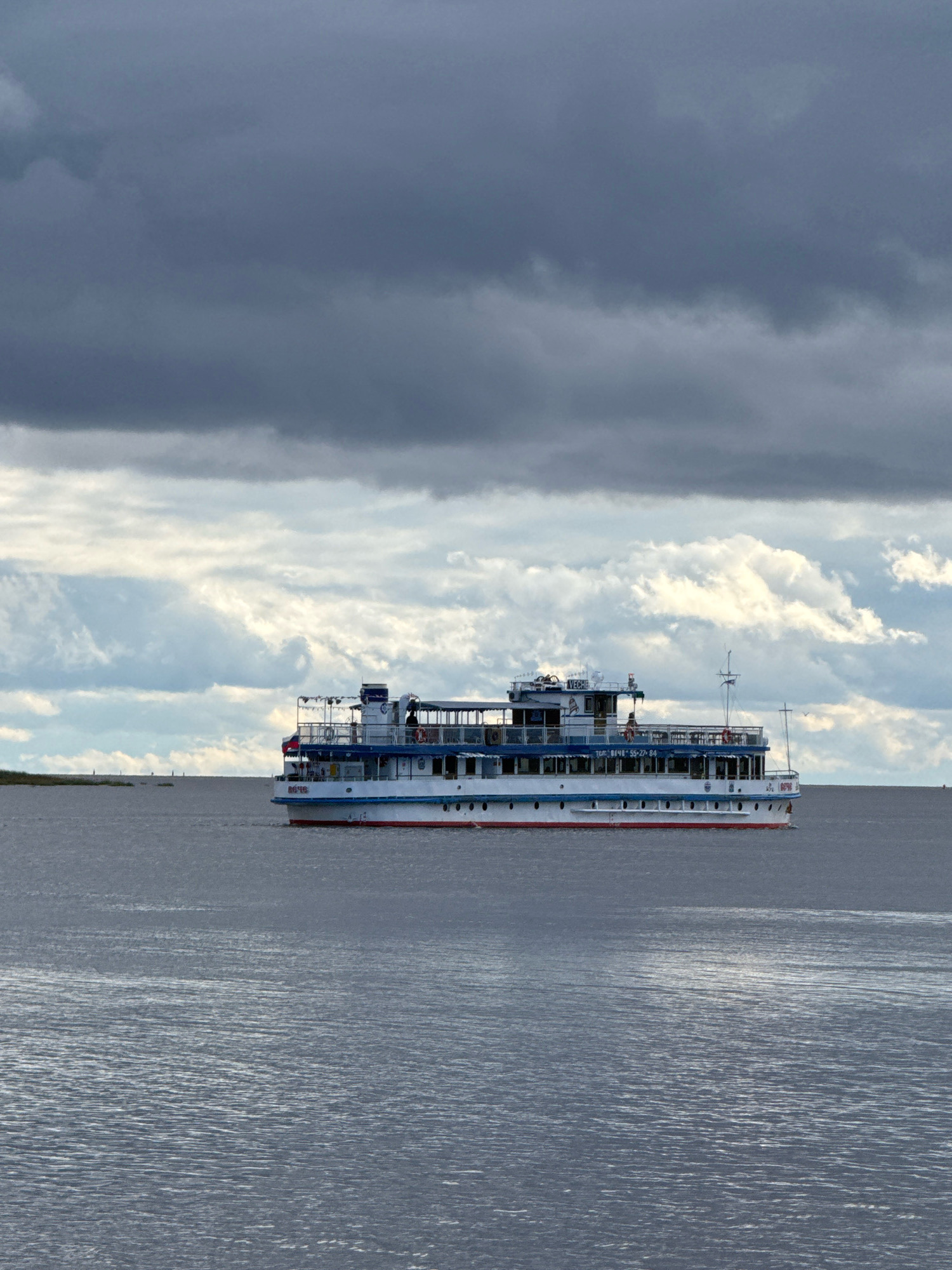 A large passenger ship, likely a riverboat, is sailing on a body of water under a cloudy sky. The ship is white with blue accents and a red waterline. It has multiple decks with railings and windows. The ship's name, "ВЕЧЕ," is visible on its side, along with a phone number. The water is choppy and reflects the sky. In the distance, a faint landmass with some structures can be seen. The sky is filled with dramatic, dark clouds interspersed with lighter, brighter clouds. The scene is likely a view of the Volkhov River or Lake Ilmen in Veliky Novgorod, Russia, given the ship's name "ВЕЧЕ," which is a historical term referring to a popular assembly in ancient Rus' and is a prominent symbol of Veliky Novgorod.