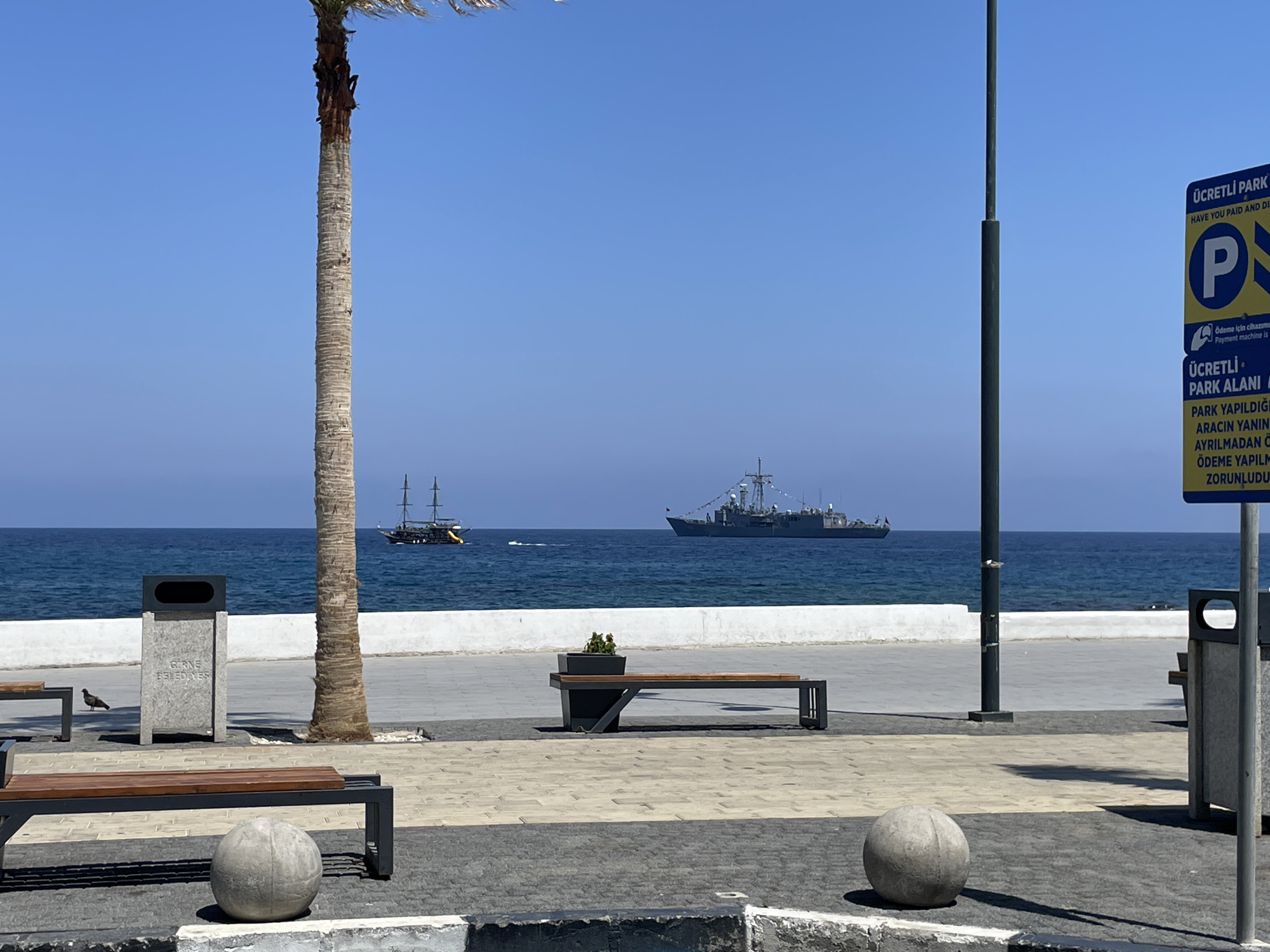 The image depicts a waterfront promenade in Kyrenia, Northern Cyprus. In the foreground, a gray stone ball rests on a paved area. To the left, a modern park bench with a wooden seat and dark metal frame is positioned. Behind it, a stone trash receptacle with the inscription "GİRNE BELEDİYESİ" (Kyrenia Municipality) is visible, with a pigeon standing near its base. A tall palm tree with a textured brown trunk dominates the left side of the frame.
In the middle ground, the promenade continues with more paved sections and benches. A dark metal bench with a wooden seat, featuring a planter box on one end, is centrally located. Beyond this, the blue sea stretches to the horizon under a clear blue sky. Two vessels are visible on the water: a sailing ship with multiple masts to the left, and a modern naval ship to the right.
On the right side of the image, a tall gray pole stands, and next to it is a yellow and blue parking sign with Turkish and English text indicating "ÜCRETLİ PARK ALANI" (Paid Parking Area) and "HAVE YOU PAID AND DISPLAYED" with a large blue "P" symbol. Another similar trash receptacle is partially visible on the far right. The overall scene is one of a public space by the sea on a sunny day.