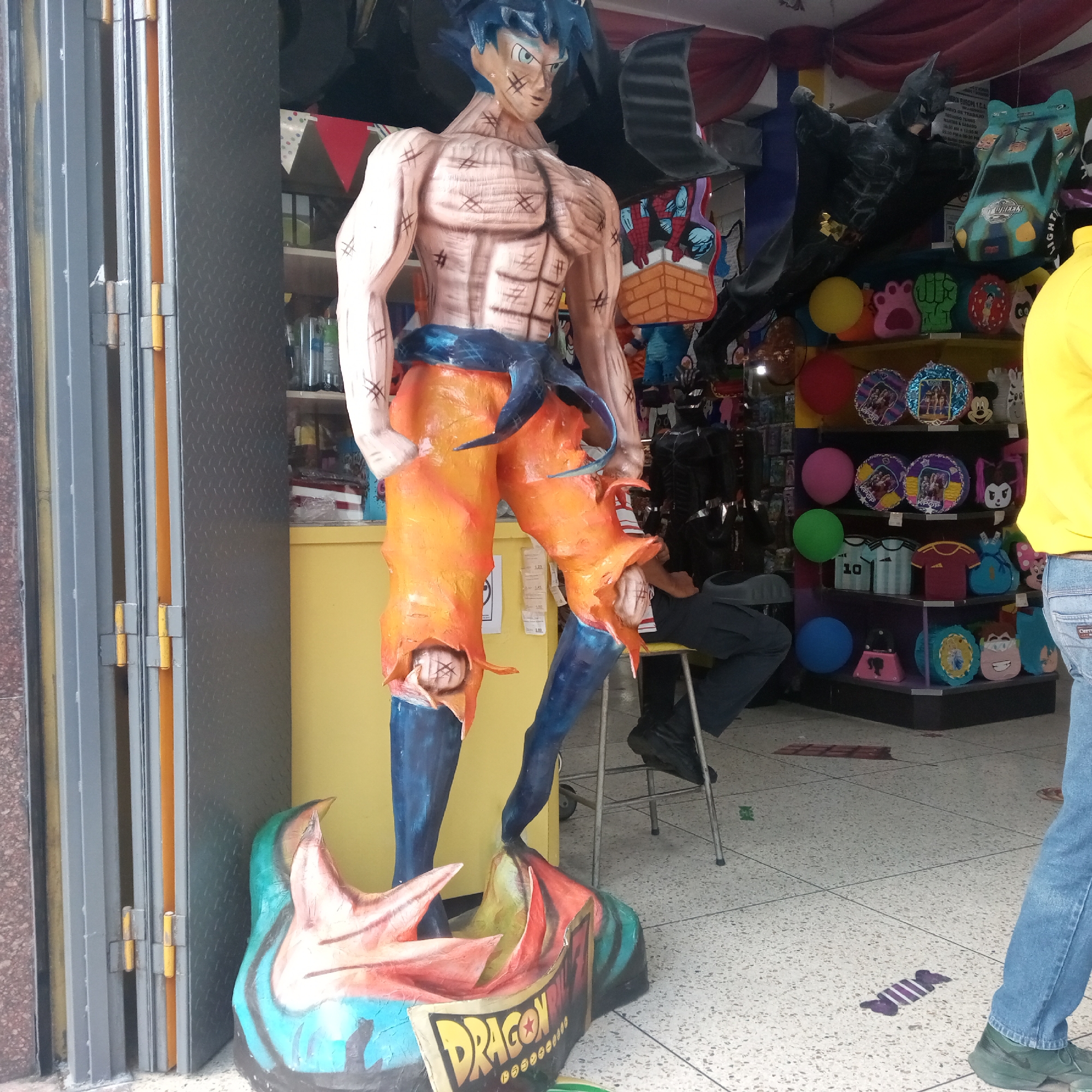 The image shows a life-size, somewhat crudely crafted statue of a muscular Dragon Ball Z character, possibly Goku, standing in the entrance of a store. The statue is painted with orange pants, a blue belt, and a blue wig. It stands on a base with the word "DRAGON" on it. Behind the statue, the interior of the store is visible, packed with merchandise, including piñatas of various cartoon characters such as Batman, cars, and other colorful party supplies. A person is sitting on a metal stool inside the shop, partially obscured by the statue. Another person, partially visible on the right edge of the image, is wearing a yellow shirt and blue jeans. The entrance has a metal gate. The floor is tiled, and there is a sticker resembling a wrapped candy on the floor. Based on the available information, it is likely the location is in Maracay, Venezuela.