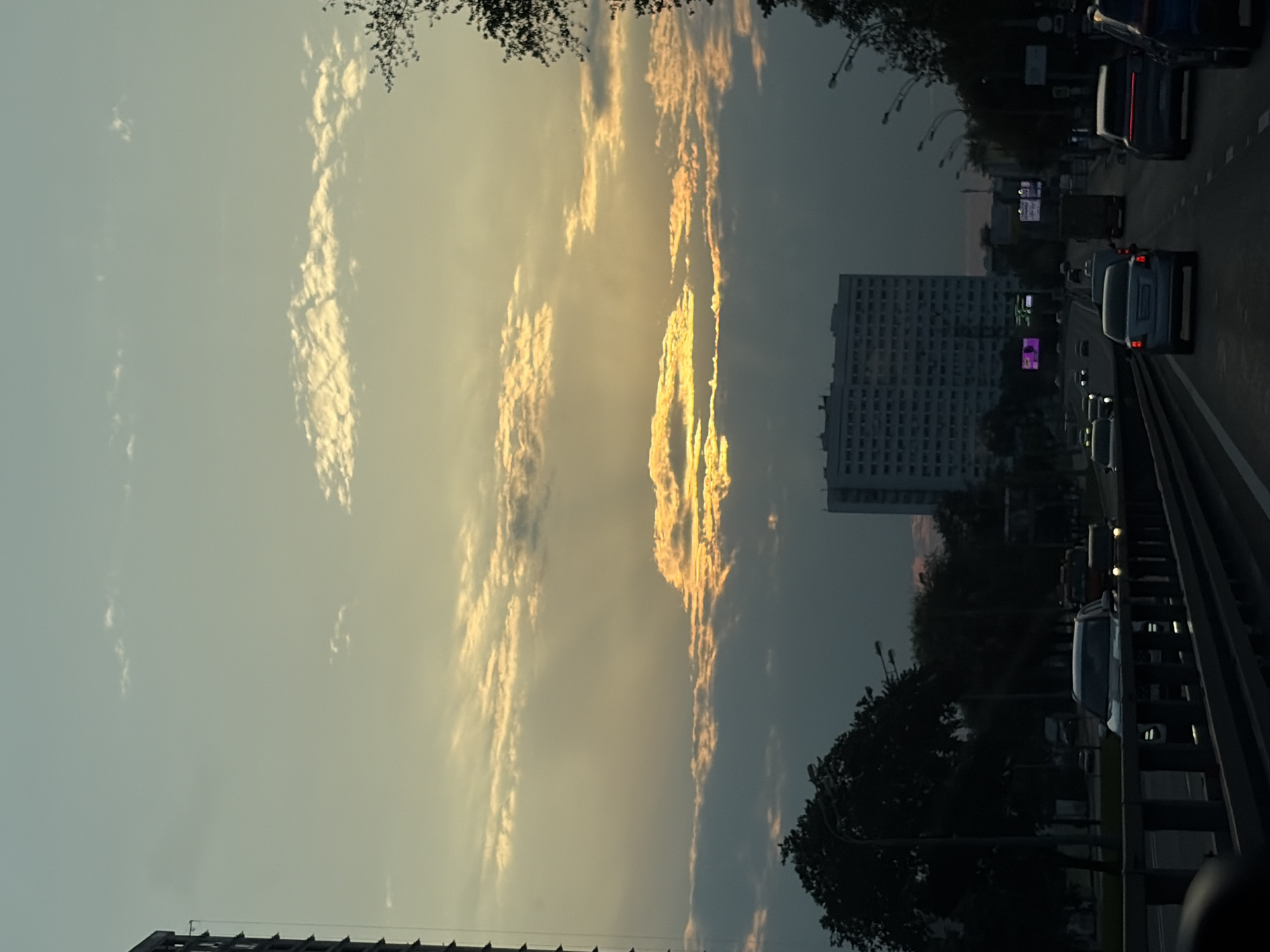 The image captures a twilight scene on a multi-lane road in Moscow, Russia. The sky is filled with a gradient of colors from pale yellow to dusky blue, with dramatic clouds illuminated by the setting sun. Sunbeams pierce through the clouds, creating a striking visual effect.
In the foreground, a bridge railing is visible, suggesting the vantage point is from a vehicle or elevated walkway. The road itself is busy with traffic, featuring various cars and trucks moving in the same direction. Among them, a white SUV is prominently displayed in the middle lane. The road is lined with trees, some of which are silhouetted against the sky.
Further down the road, a large, light-colored residential building stands tall, characteristic of Moscow's urban landscape. Streetlights and traffic signals are also present, indicating the urban setting. The overall atmosphere is one of an active city transitioning from day to night.