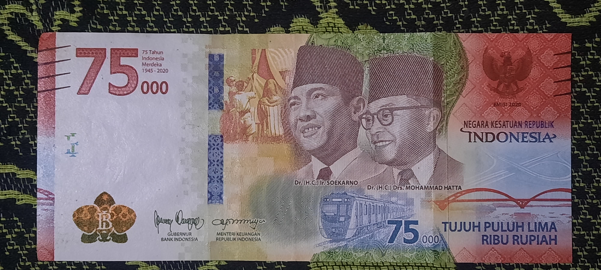 The image is a close-up, top-down view of a 75,000 Indonesian Rupiah banknote. The banknote is laid flat on a textured dark green fabric. The banknote features portraits of two men, identified as Dr. (H.C.) Ir. SOEKARNO and Dr. (H.C.) Drs. MOHAMMAD HATTA, wearing traditional caps. The number "75" is prominently displayed on the left side of the note, along with text indicating "75 Tahun Indonesia Merdeka 1945-2020". A stylized orchid with the letter "B" inside it is visible on the lower left. The national emblem of Indonesia, Garuda Pancasila, is on the upper right, with the text "NEGARA KESATUAN REPUBLIK INDONESIA" below it. The value of the banknote is also written in words as "TUJUH PULUH LIMA RIBU RUPIAH". A depiction of a modern train and a bridge are also part of the design. The banknote is in good condition. There is no discernible location in Jatitengah, Indonesia visible in this image.