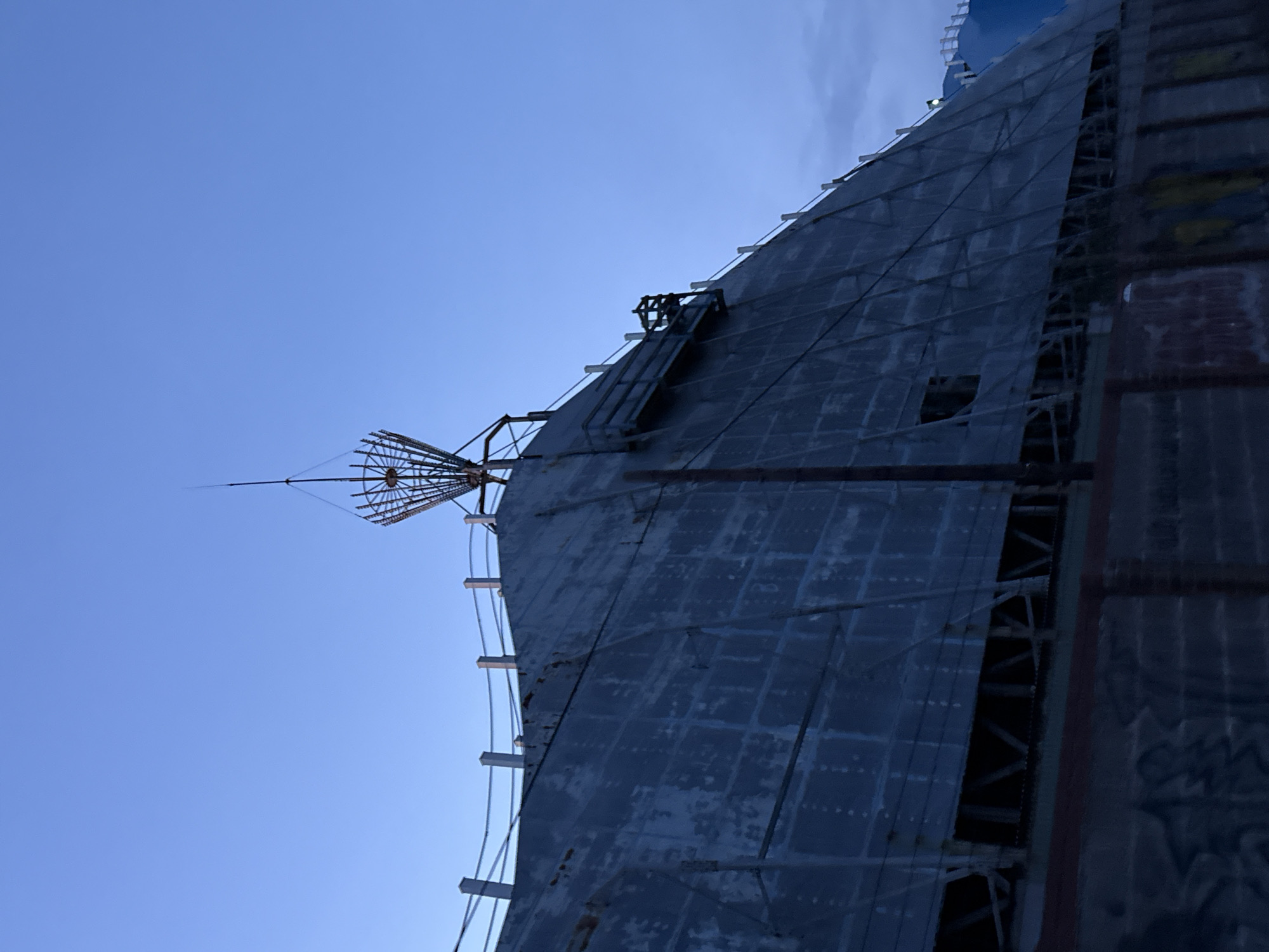 The image shows the Gorleston-on-Sea Transmitter, a large radio tower structure in Gorleston-on-Sea, United Kingdom. The tower has a metal skin composed of grid-like panels, and a visible antenna at the top. There are ladders and scaffolding attached to the side. The sky is mostly clear with blue hues.