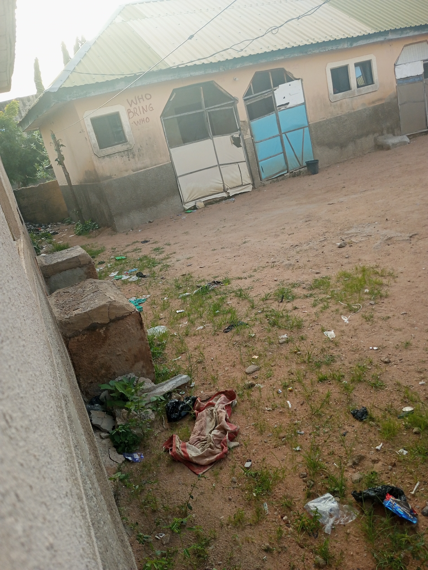 The image depicts an outdoor scene in Jalingo, Nigeria, showing the exterior of a building with a light yellow facade and a corrugated metal roof. The building has at least two distinct entrances with metal framing and panels, one of which is partially covered with a white material and the other with light blue panels. A window with dark panes is visible on the upper part of the building, and graffiti reading "WHO BRING WHO" is present on the facade.
The ground in front of the building is unpaved, sandy, and sparsely covered with grass and weeds. There is evidence of litter scattered across the ground, including plastic bags, wrappers, and some cloth items. Specifically, a folded cloth with red and white stripes is visible on the ground. Two large concrete blocks are positioned near a wall on the left side of the image, and further debris and vegetation are present around them. A small black bucket is situated near one of the building's entrances. The overall impression is of an outdoor area that is somewhat neglected and unkempt. The lighting suggests it is daytime, with bright sunlight casting shadows.