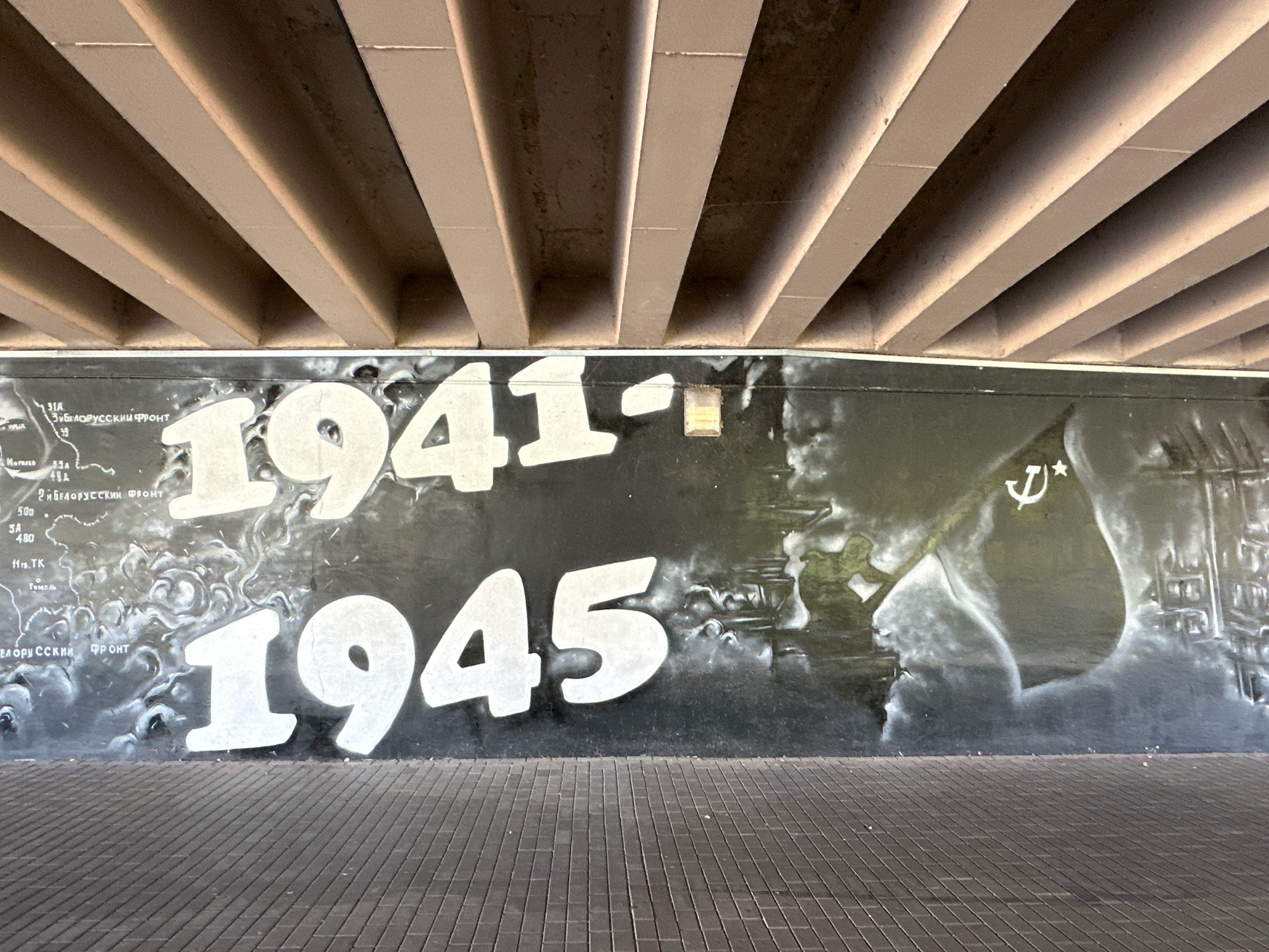 The image depicts a large mural painted on a wall beneath an elevated structure, likely a bridge or overpass. The mural features the years "1941" and "1945" in large, white, stylized numbers. Between these dates, the mural contains a depiction of a map with Cyrillic text, as well as imagery that appears to represent warfare, including figures, explosions, and a flag with a star and hammer and sickle symbol. The text "31K ЗИБЕЛОРУССКИЙ ФРОНТ" (31st Belorussian Front) is visible on the map. The ground in front of the mural is paved with dark gray rectangular tiles. The overall style is a combination of graphic lettering and airbrushed imagery in black and white tones. The mural is located on the embankment of the Western Bug river in Brest, Belarus, commemorating events related to World War II.