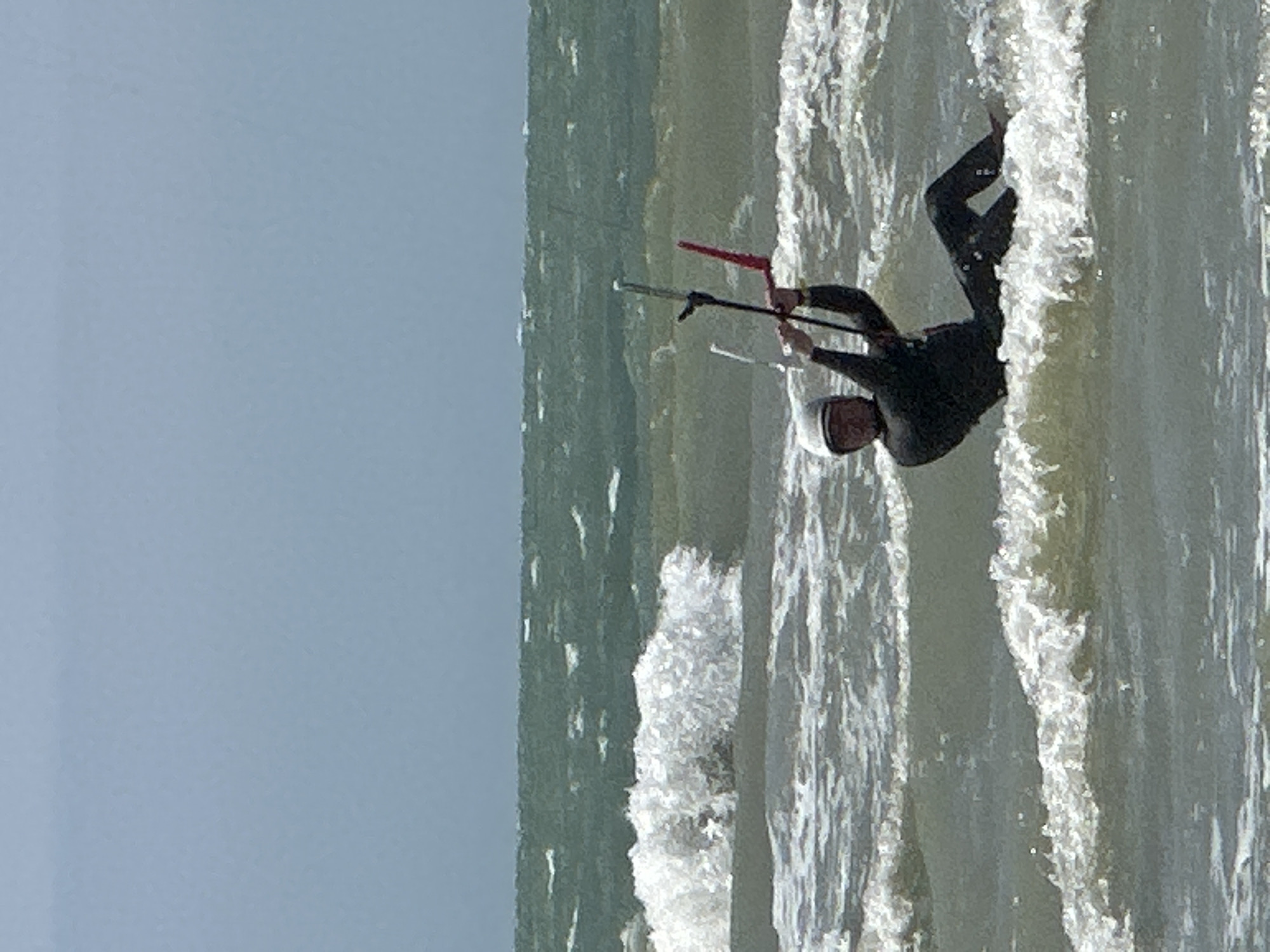 The image captures a kitesurfer in the water off the coast of Berck, France. The kitesurfer, wearing a black wetsuit and a white helmet, is leaning back, controlling the kite with a bar. They are partially submerged in choppy, white-capped waves. The sea is a murky green, with the horizon line of a pale blue sky visible in the background. The scene conveys an active, outdoor sport in a coastal environment.