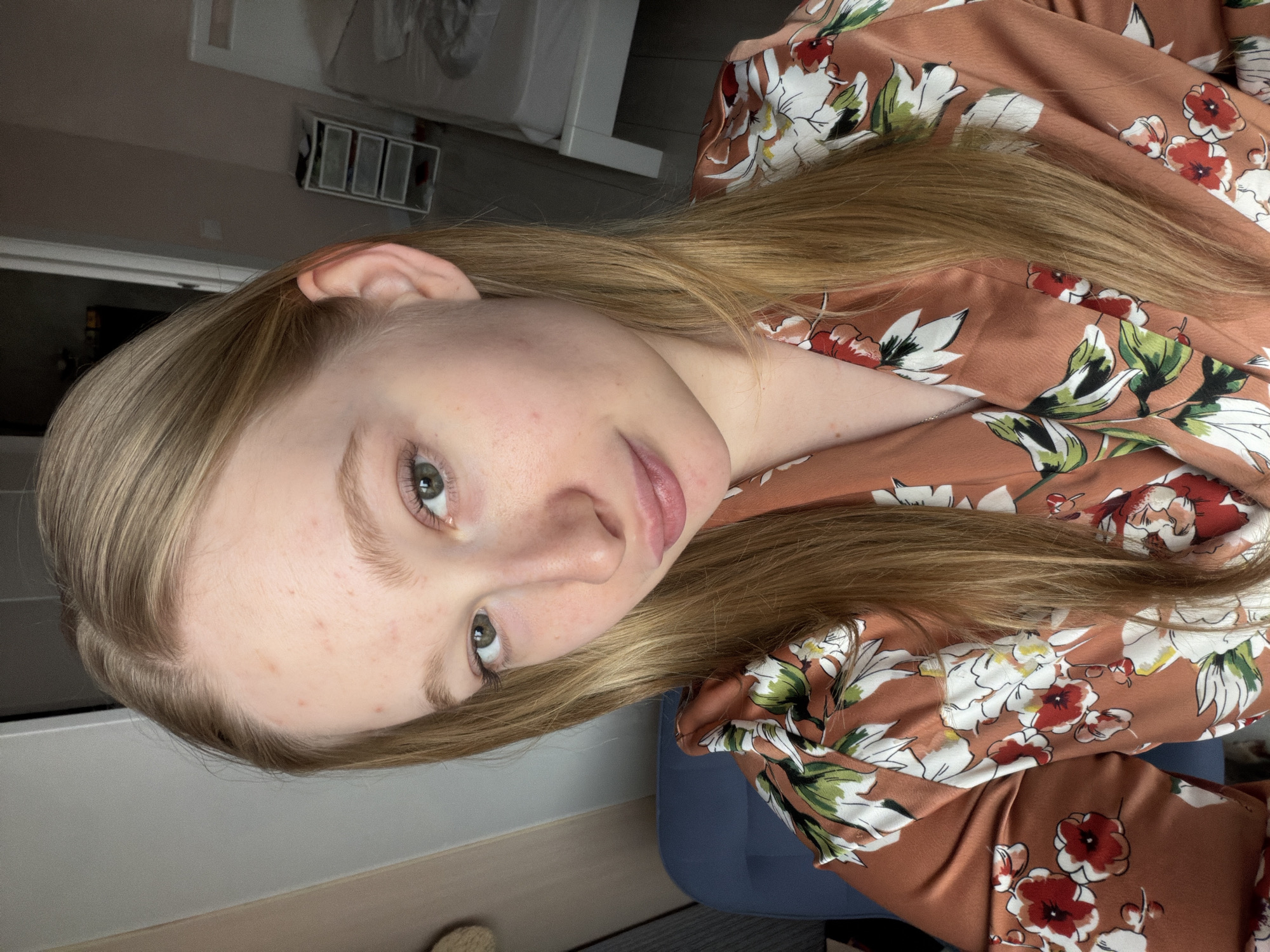 A young, light-skinned person with long blonde hair and green eyes is facing the camera. They are wearing a floral-patterned shirt in shades of brown, red, white, and green. The person has visible skin blemishes. The setting appears to be indoors. The location in Rossiyskiy, Russia, is not discernible from the image.