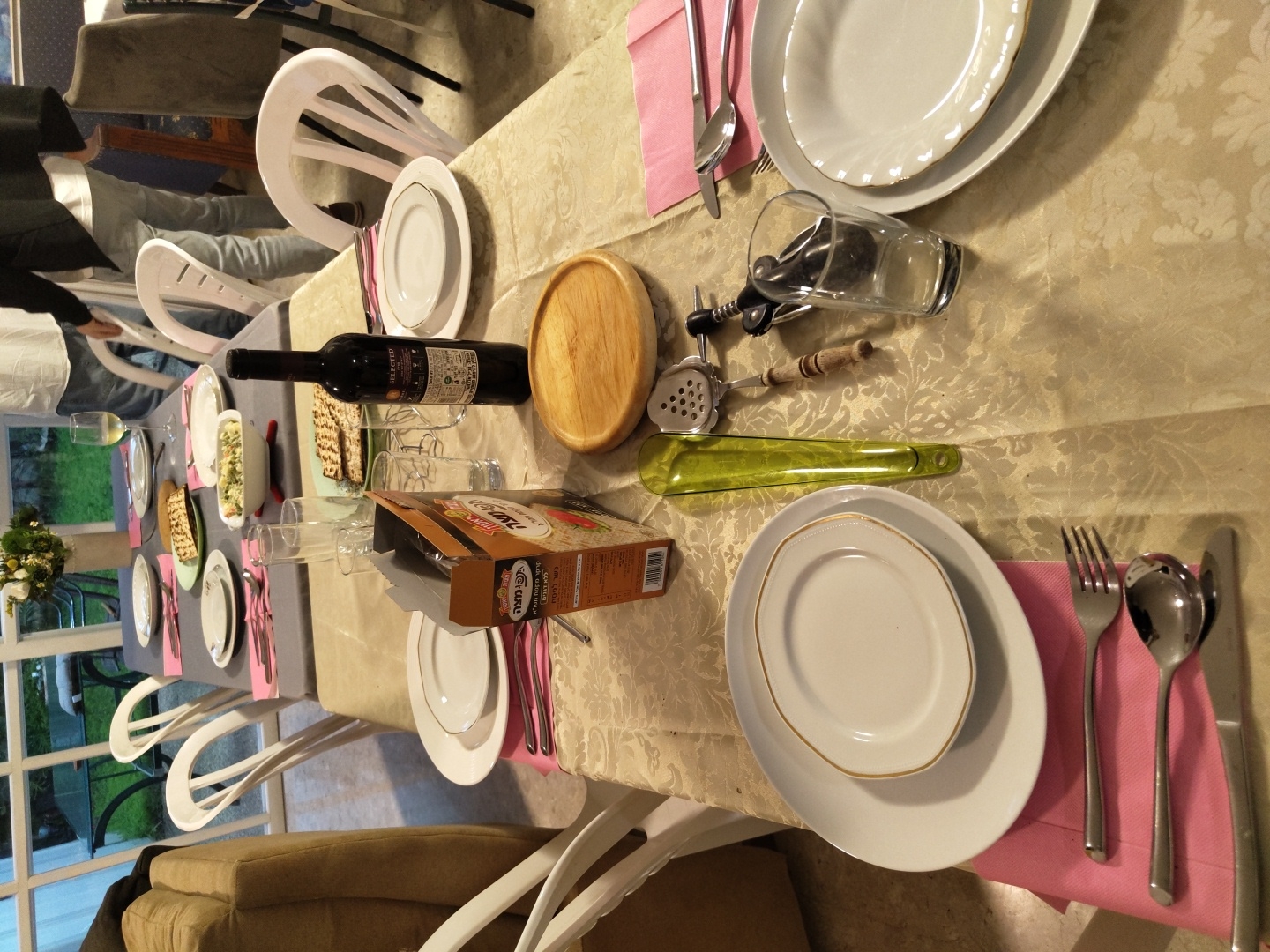 The image shows a dining table set for a meal, likely indoors. There are multiple place settings with plates, cutlery, napkins, and glasses. A bottle of wine and a box of matzah are on the table. A wooden serving platter, wine opener, and other serving utensils are also present. There are chairs around the table, some of which are occupied by people. A window in the background shows an outdoor area. Based on the box of matzah and the presence of a Seder plate, the meal may be for Passover. According to the information included with the image file, the location is Raanana, Israel.
