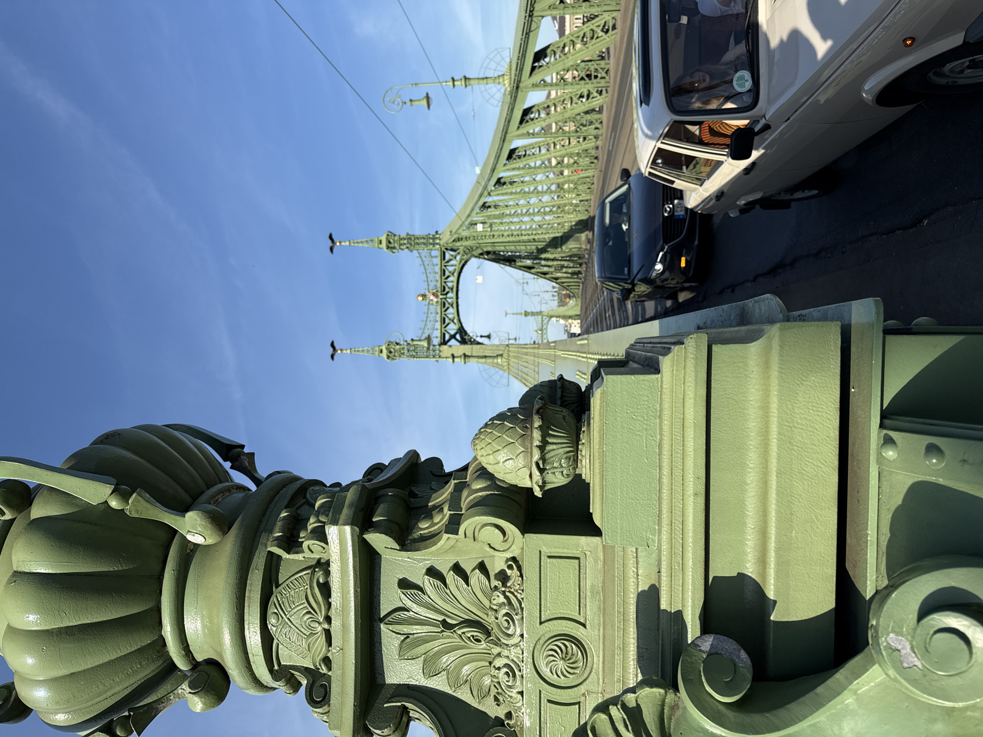 The image depicts the Liberty Bridge in Budapest, Hungary, viewed from a low, upward angle. A section of the bridge's green metalwork is in the foreground, featuring decorative floral and structural elements. Beyond, the bridge's steel structure stretches into the frame, with the bright blue sky visible through its arches. Two vehicles, a black car and a white van, are partially visible on the bridge's roadway.