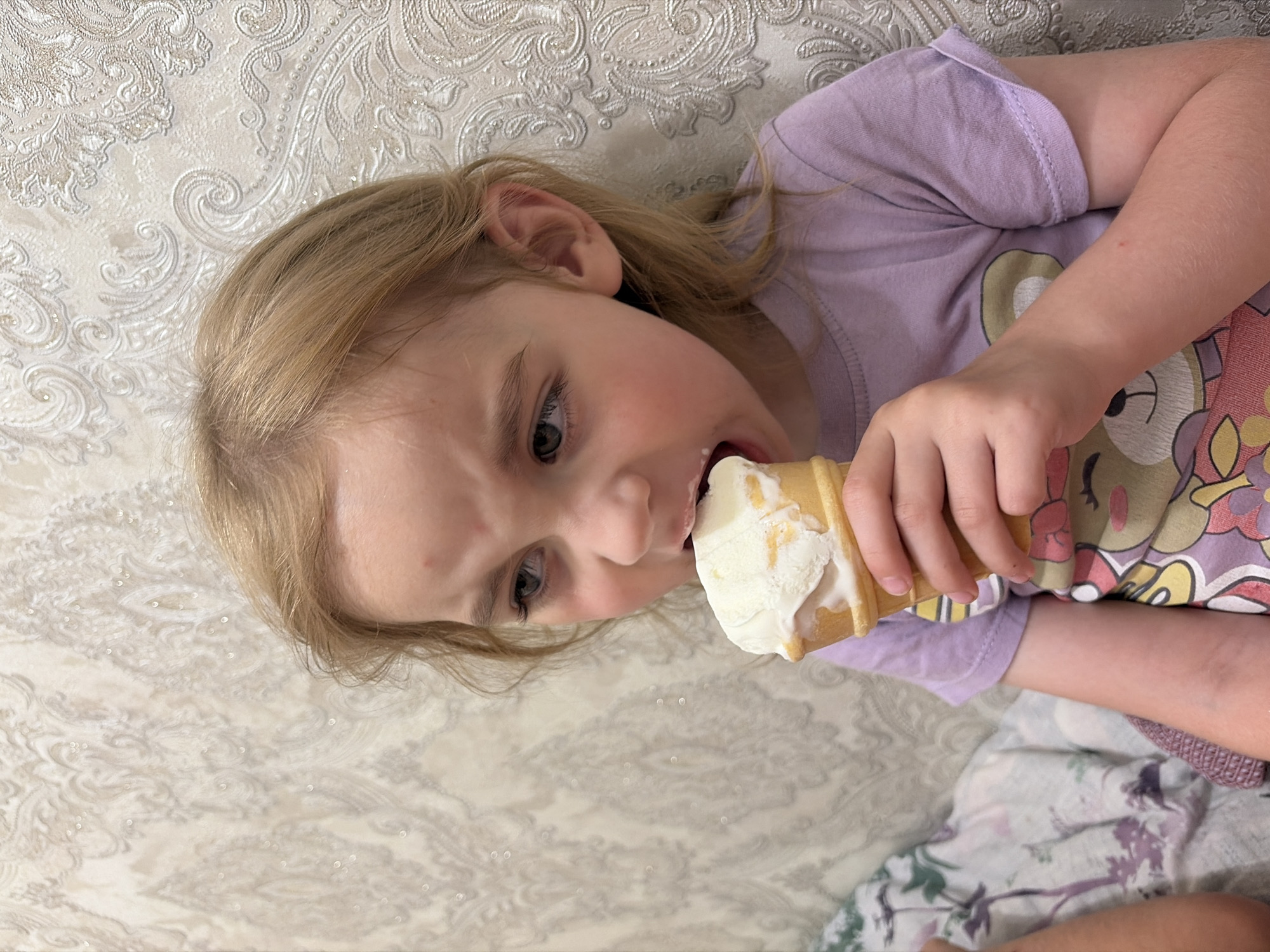 A young blonde girl is sitting and eating an ice cream cone. She is wearing a light purple t-shirt with a cartoon bear on it. The background is a patterned wallpaper. The image does not provide any information about the location in the city of Tambov, Russia.