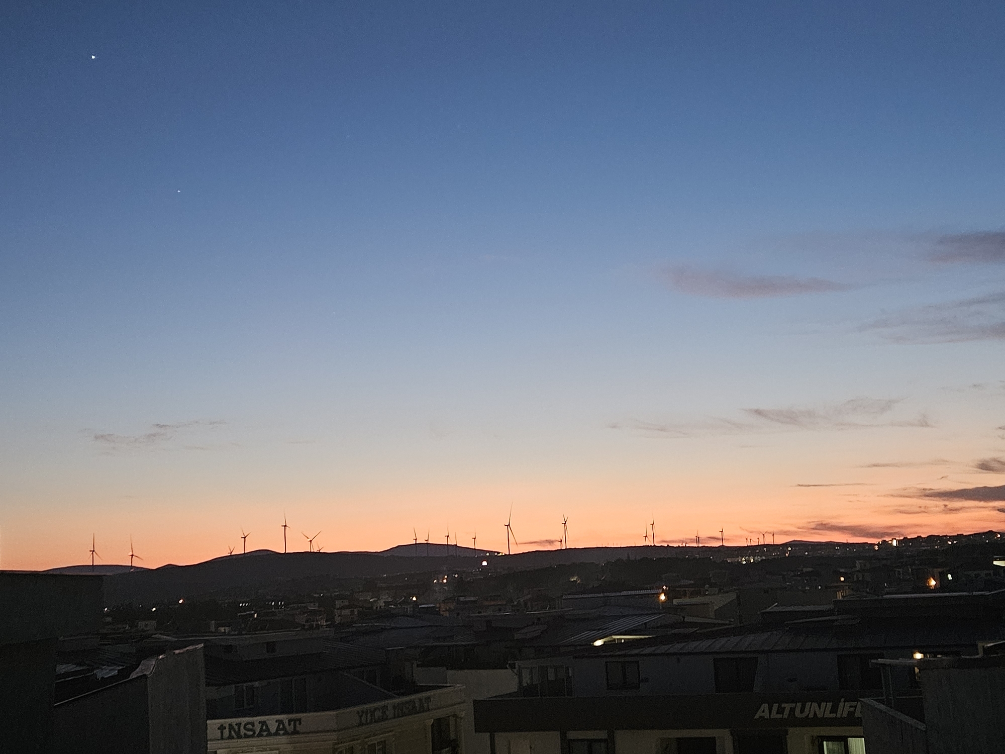 The image shows a cityscape at dusk with a clear sky transitioning from deep blue to orange. Wind turbines are visible on a hilly horizon in the distance. In the foreground, several buildings are silhouetted against the sky, some with visible signage. The overall scene depicts a twilight view of Seferihisar, Turkey, characterized by urban development and renewable energy infrastructure.
