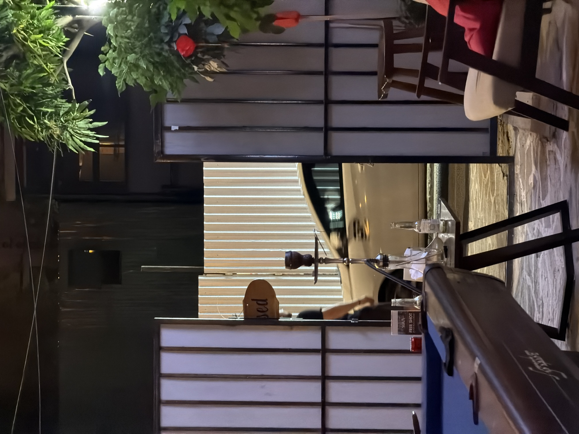 This media file captures a nighttime scene at what appears to be an outdoor or semi-outdoor bar or lounge in Đà Nẵng, Vietnam. The image is oriented vertically but depicts a horizontal view.
In the foreground, a dark, long object, possibly a pool table or a large counter, extends across the bottom left. On an adjacent counter or table, several clear glass bottles are visible, one with a pourer, along with a small white sign. Prominently positioned in the mid-ground is a tall, ornate hookah (shisha pipe). Behind it, a person's arm and head are partially visible, seemingly engaged in preparing or serving something behind a counter made of light-colored horizontal slats. A wooden sign, partially obscured, shows the text "sed," likely part of "closed."
The setting is characterized by lush green foliage hanging overhead, interspersed with a few artificial red flowers, suggesting a vibrant, natural ambiance. Bright artificial lighting illuminates the scene from above the greenery. In the background, beyond an opening in the structure, a white vehicle (possibly a limousine or van) is parked on a street next to a corrugated metal wall or garage door. To the far right, wooden chairs with cushioned seats, one with a red cushion, are arranged against a stone or textured wall.
The activity suggests a relaxed social gathering. The visible text includes "SWAIN FREE SHOT" on a small white sign and partially "Jacc" on the side of the dark foreground object, possibly a brand name. The dark surroundings and artificial lights clearly indicate that the photograph was taken at night.