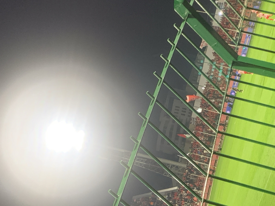 A nighttime stadium view through a green metal fence. Bright stadium lights glare, illuminating a sports field. A multi-tiered stadium with many spectators is visible in the background. Flags and banners can be seen among the crowd. The sky is dark, suggesting a night game. The fence and bright light are in the foreground, contrasting with the activity in the stadium.