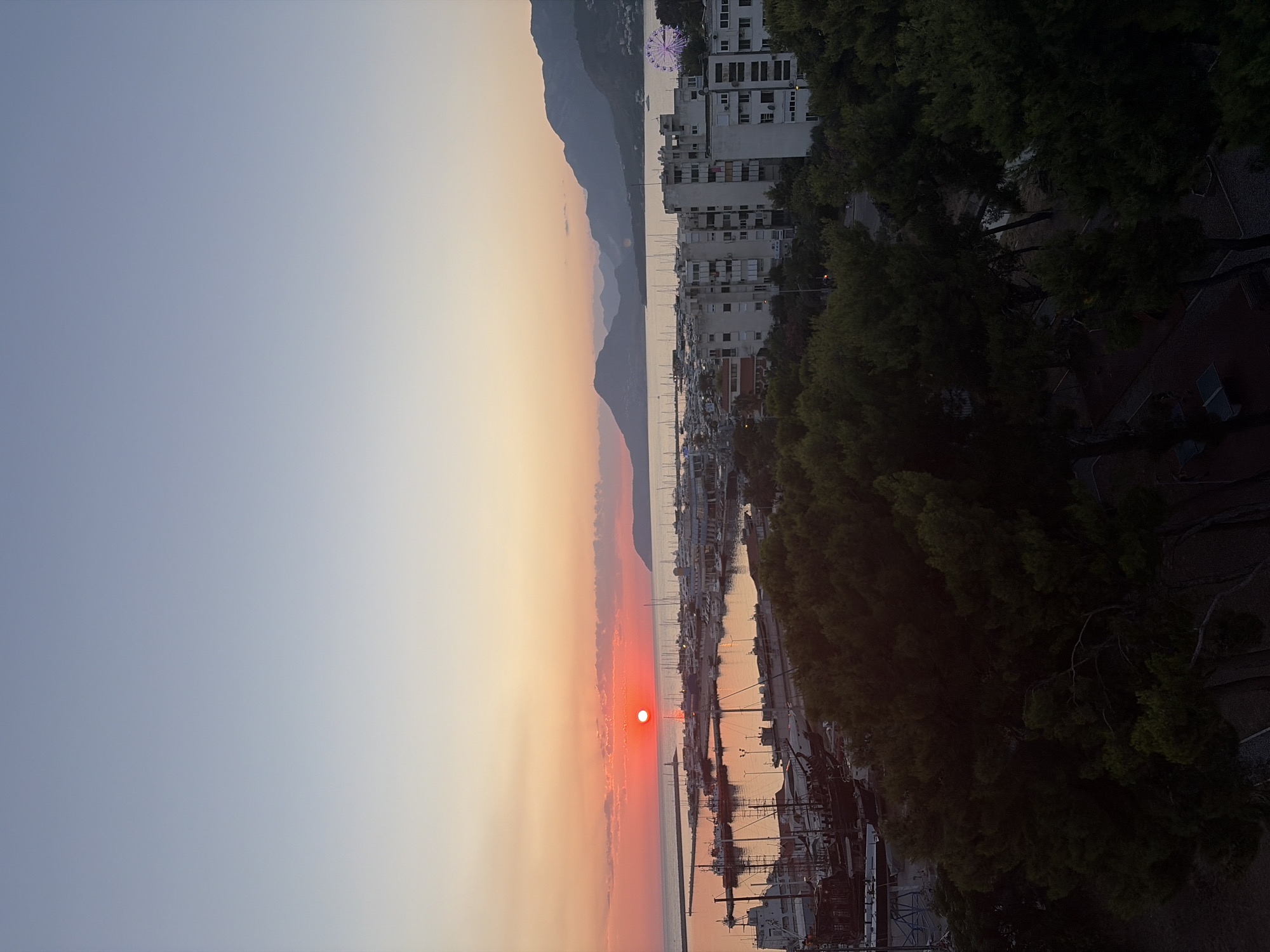 The image captures a sunset over a harbor in Bar, Montenegro. The sun, a vibrant orange orb, is positioned low on the horizon, casting a warm glow across the sky and water. The sky transitions from a pale yellow near the sun to a soft blue higher up. Clouds are scattered across the horizon, tinged with the sunset's colors.
The harbor is filled with numerous boats, including sailboats and larger vessels, docked in neat rows. A lighthouse stands at the entrance to the harbor. Beyond the harbor, a city skyline is visible, characterized by apartment buildings. In the background, a Ferris wheel with pink lights is illuminated, suggesting it is in operation. Further in the distance, rolling mountains form a picturesque backdrop. In the foreground, lush green trees fill the bottom portion of the frame, obscuring some of the lower structures and parts of the city. The overall scene depicts a peaceful coastal evening.
