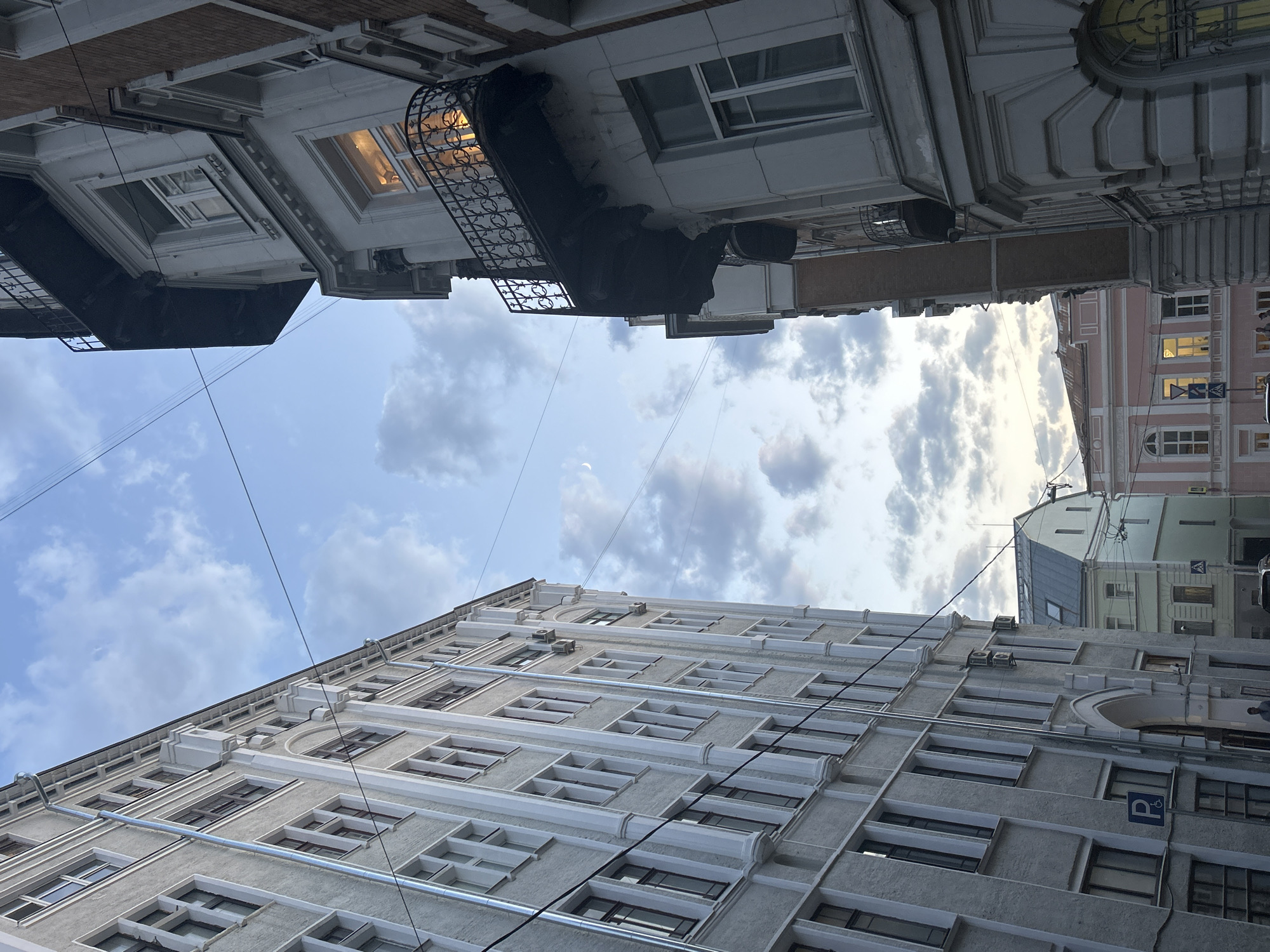 The image depicts a narrow street view in an urban environment, likely in the city of Unknown, Unknown. The photograph is taken from a low angle, looking upwards towards the sky, which is partially filled with clouds.
Two buildings frame the street. One building on the lower portion of the image is a tall structure with multiple floors. It has a grey facade with numerous rectangular windows. The windows are framed with white trim and have dark window frames. There are also drainpipes running vertically down the building's exterior.
The second building is on the upper portion of the image and appears to be lighter in color, possibly white or off-white. It has decorative architectural details, including window frames and a wrought iron balcony. Some windows are illuminated from the inside.
Power lines run across the sky, intersecting the view of the buildings and the sky.
The overall impression is that of an old European city, with classic architecture. The lighting indicates that the photograph was taken either during the day or at twilight. There are no visible people in the frame.