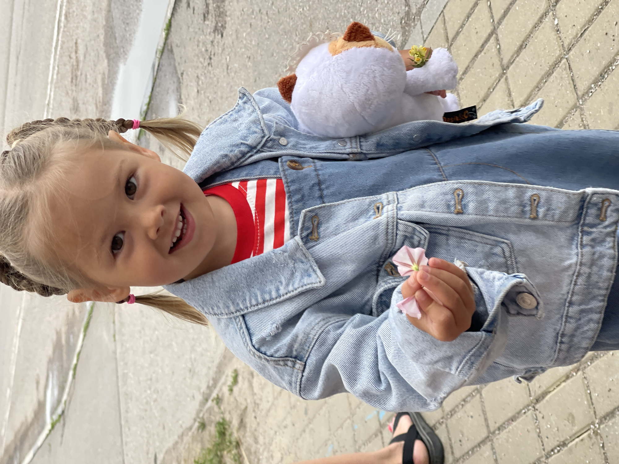 A young Caucasian girl with blonde hair styled in two braids, each tied with a pink elastic band, stands outdoors. She is wearing a red and white striped t-shirt, denim overalls, and a light blue denim jacket. In her right hand, she holds a small pink flower. In her left arm, she cradles a white stuffed animal with brown ears and accents. The ground is paved with light-colored rectangular stones, and a wet, grey surface, possibly a road or sidewalk, is visible in the background. The scene appears to be a street in Zelenogradsk, Russia, given the context of the request.