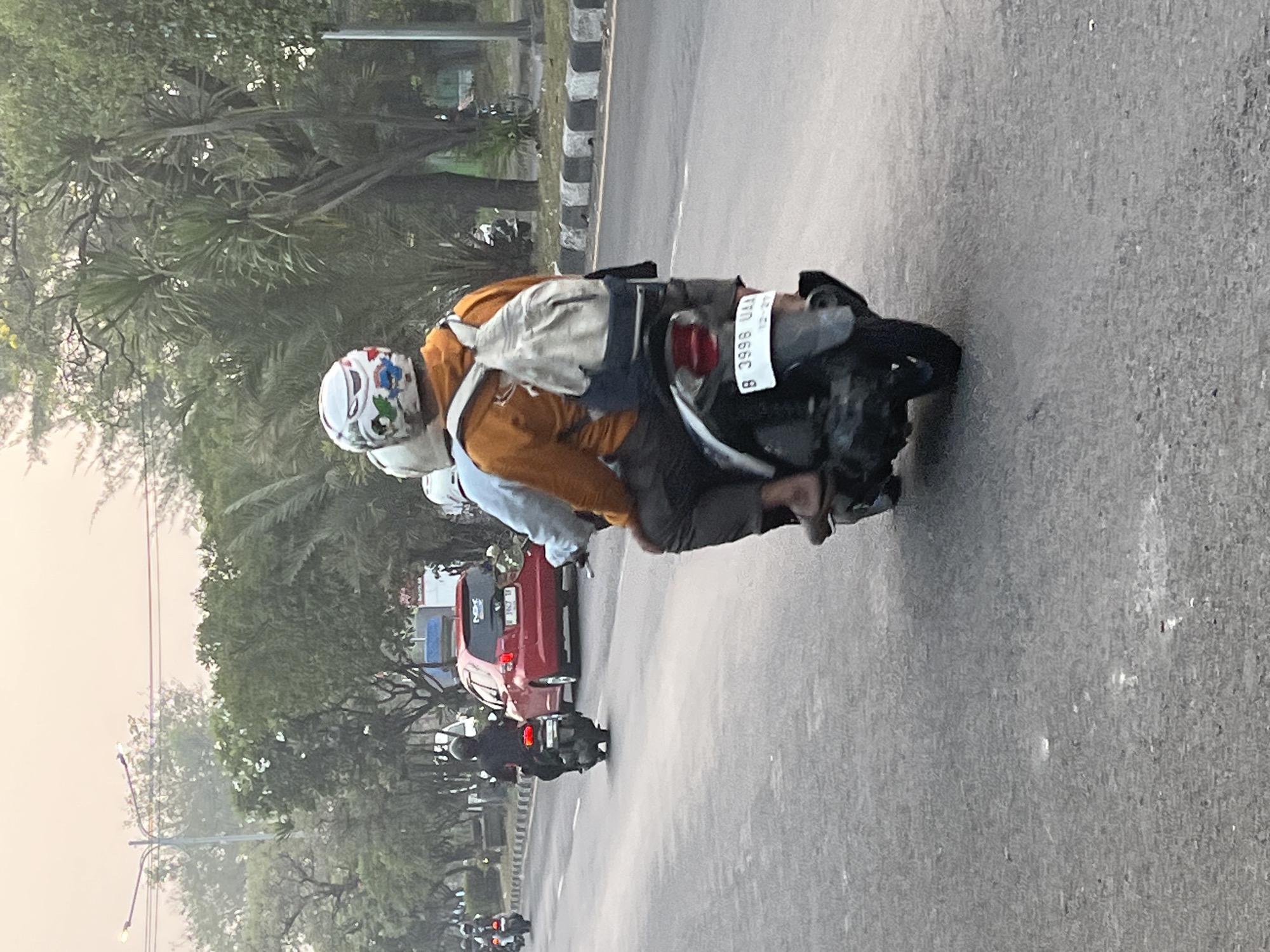 The image depicts a street scene in Ngaban, Indonesia. In the foreground, two individuals are riding on a scooter. The person in the front is wearing an orange hoodie and a backpack, while the person in the back is wearing a dark shirt and a helmet. The scooter has a license plate that reads "B 3998 UAA".
In the middle ground, a red car is driving on the road. Several other scooters and a car can be seen in the background, suggesting moderate traffic. The street is lined with trees and there are streetlights visible. The sky is overcast with a soft, diffused light, indicating it might be dawn or dusk. The overall atmosphere suggests an urban environment.