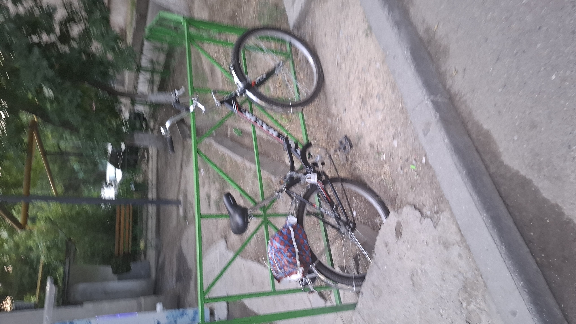 Two bicycles are parked in a green metal rack. The rack is situated outdoors, next to a concrete curb and a paved area. Behind the rack, there are trees and foliage. One bicycle is black with red and white accents on its frame. It has a black saddle and a bag with a red and blue checkered pattern is hanging from its handlebars. The other bicycle, partially visible, appears to be silver or grey. The scene suggests an outdoor public space, possibly a park or a street in Shymkent, Kazakhstan. The lighting indicates it is daytime, and the weather appears to be fair. There is no visible text in the image.