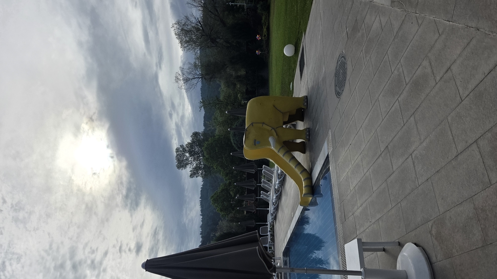 An outdoor swimming pool area is depicted under a cloudy sky with the sun partially visible through the clouds. In the foreground, a blue swimming pool is to the left, with a tiled walkway surrounding it. To the right of the pool, a yellow elephant-shaped slide with a curved chute is installed. The slide is designed with a cartoonish representation of an elephant's body and legs. Further back, a row of lounge chairs and closed umbrellas are arranged along the edge of the pool. Beyond the pool area, a grassy lawn is visible, with trees and a hillside covered in vegetation in the background. A white spherical object rests on the grass to the right of the elephant slide. The scene appears to be a resort or hotel amenity.