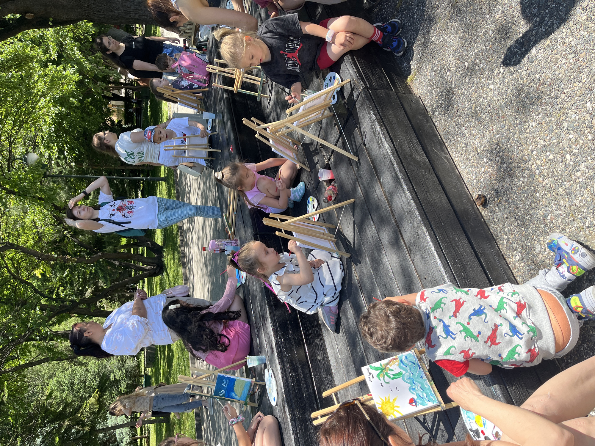 The image shows a group of children and adults participating in an outdoor painting activity. They are seated on a long, dark-stained wooden structure, likely a bench or platform. Each person has a small wooden easel with a canvas on which they are painting. Painting palettes and brushes are visible. The subjects of their paintings vary, including landscapes with suns and palm trees. The children are wearing diverse clothing, including a girl in a pink outfit, a boy in a dinosaur-patterned shirt, and a girl in a black and white dress. Some of the adults are assisting the children. The scene is set in a park with trees in the background, suggesting an urban green space. This activity may be occurring in the city of Plovdiv, Bulgaria.