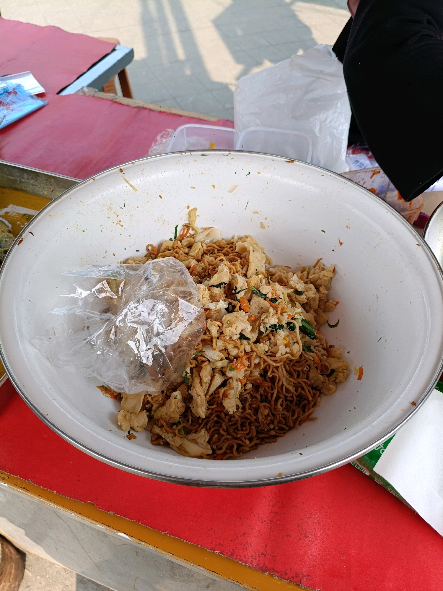 A close-up, overhead shot captures a large white bowl filled with noodles, scrambled eggs, and vegetables. The bowl is positioned on a red table, with hints of a metal tray and a yellow edge visible. A clear plastic bag containing food is placed on top of the noodle mixture, obscuring part of the contents. The background shows a blurry, outdoor environment with shadows on the ground and a white plastic bag and black fabric partially visible. The scene appears to be set in an outdoor food stall or market in Maguwoharjo, Indonesia, likely a casual dining setting.
